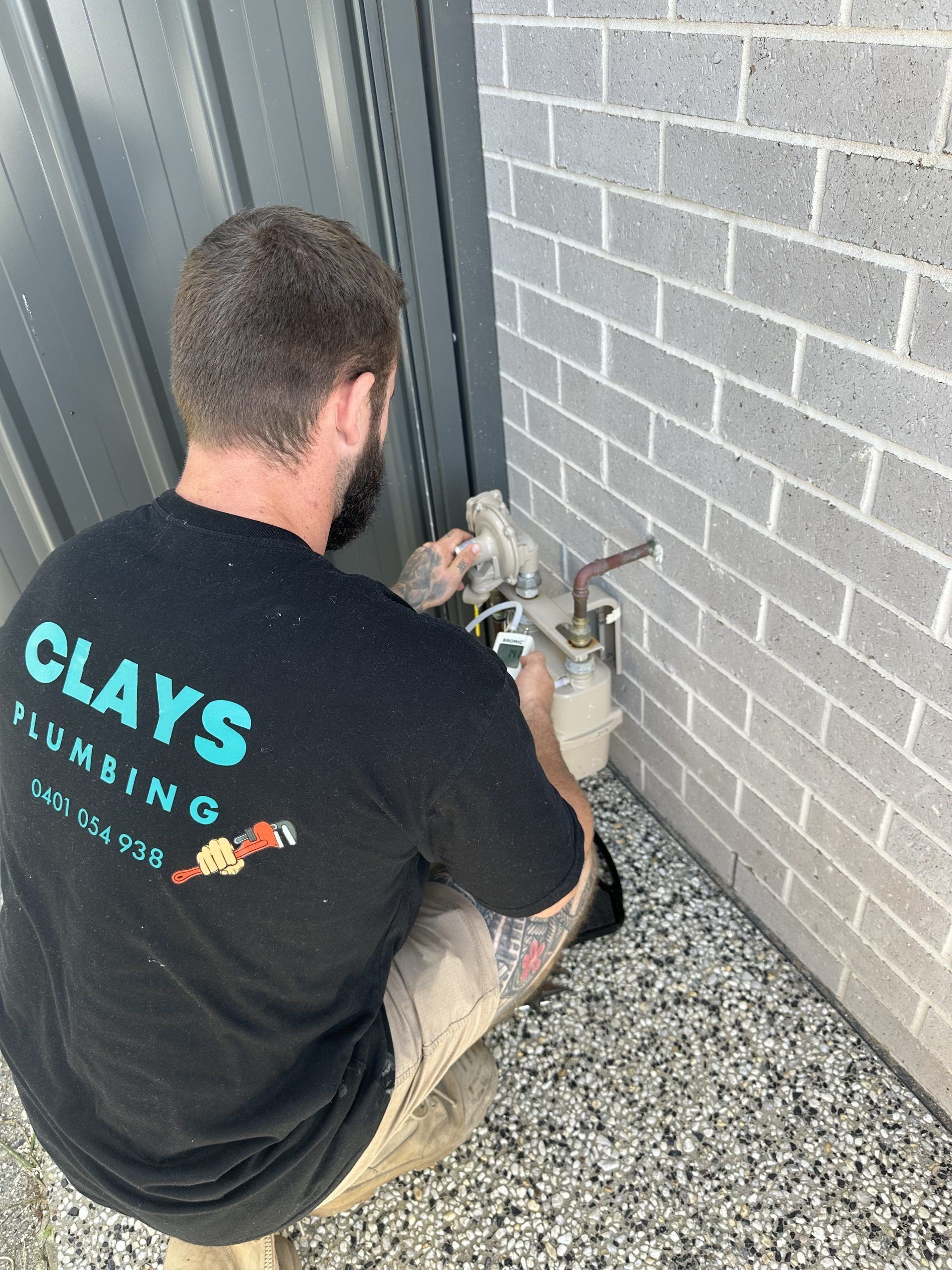 Plumber in a black shirt working on a gas meter near a brick wall and corrugated metal.