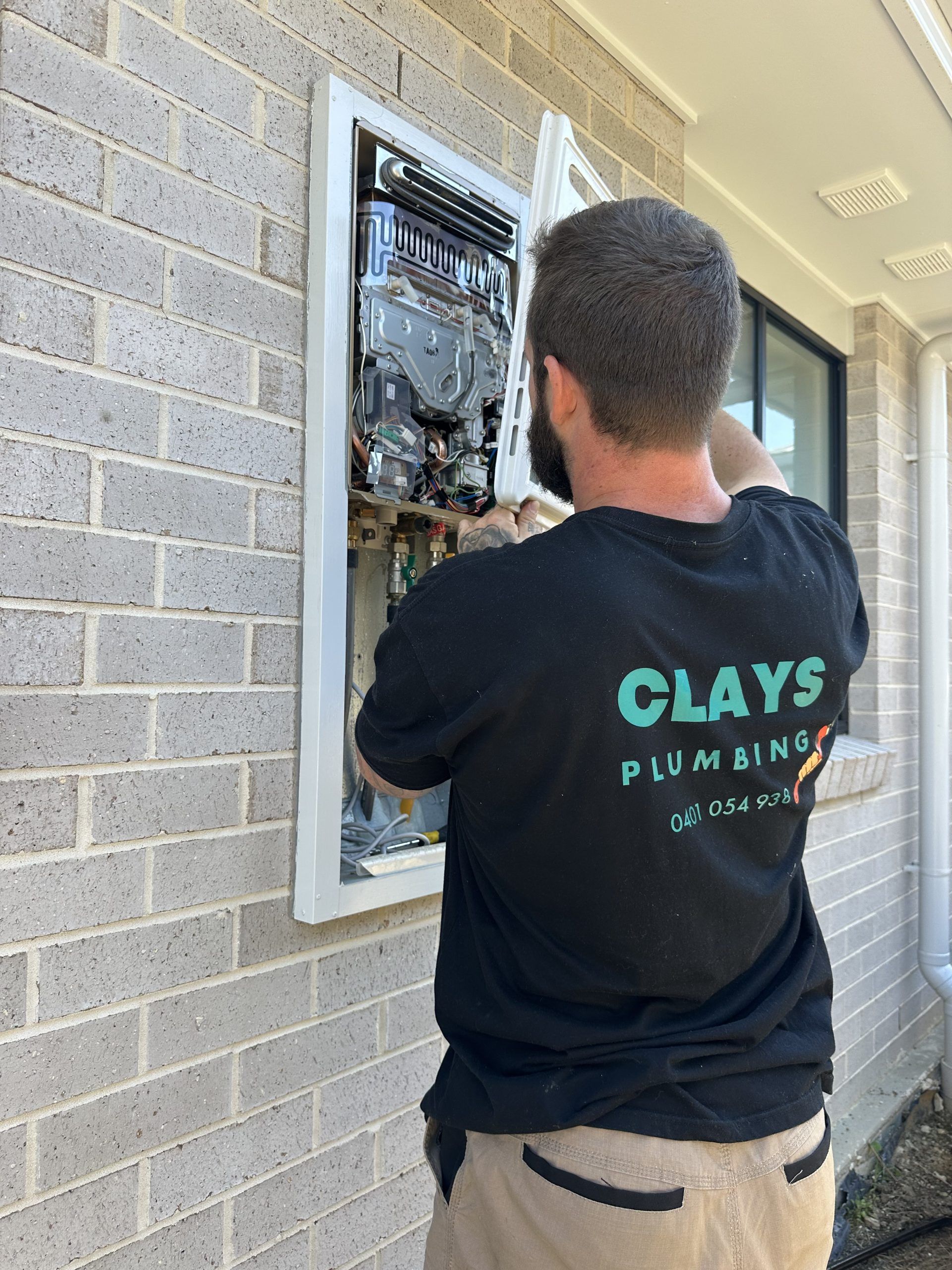 Plumber installing a water heater on a brick exterior wall. He wears a black shirt with company logo.