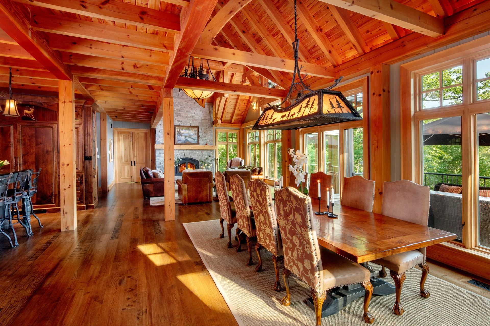 Rustic dining room with wood beams, long table, upholstered chairs, and sunlight through large windows