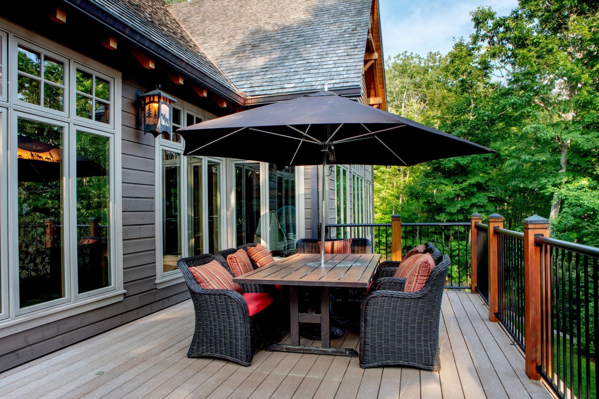 Covered patio with wicker dining set, black umbrella, and glass doors beside a wooden deck and green trees