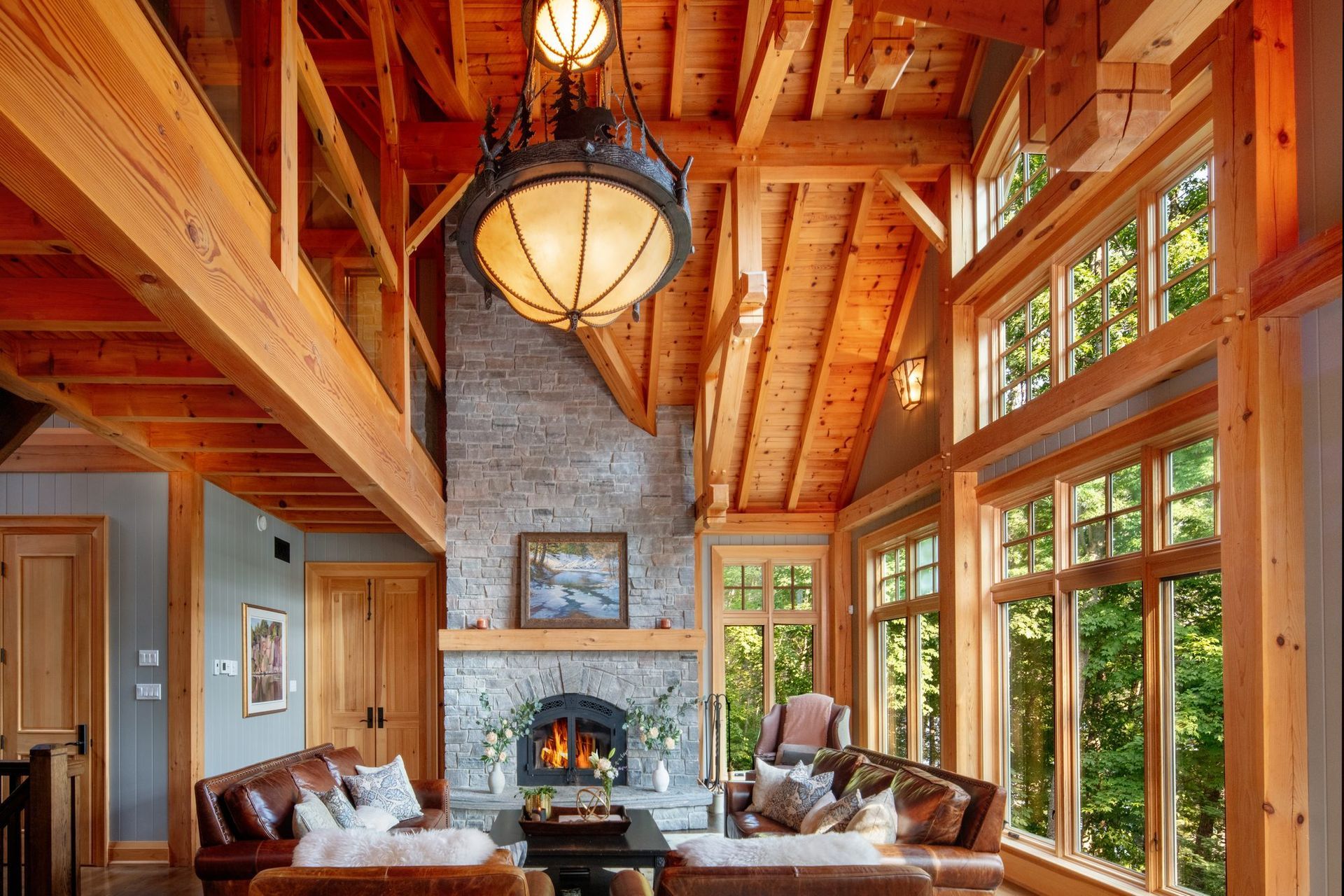 Rustic living room with exposed wood beams, stone fireplace, leather sofas, and large windows overlooking trees