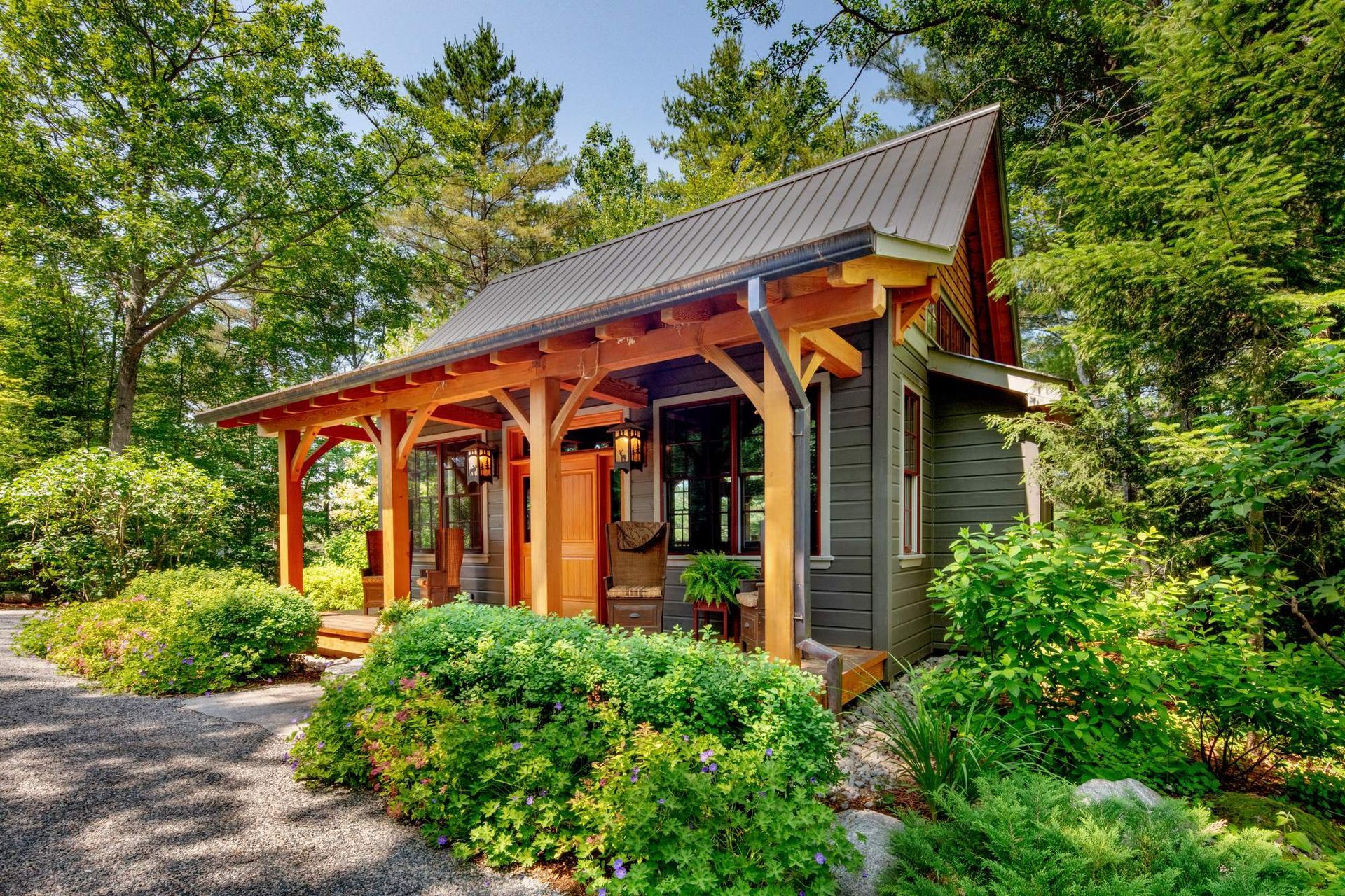 Small rustic cabin with a wooden porch, gray roof, and lush green landscaping in a wooded setting
