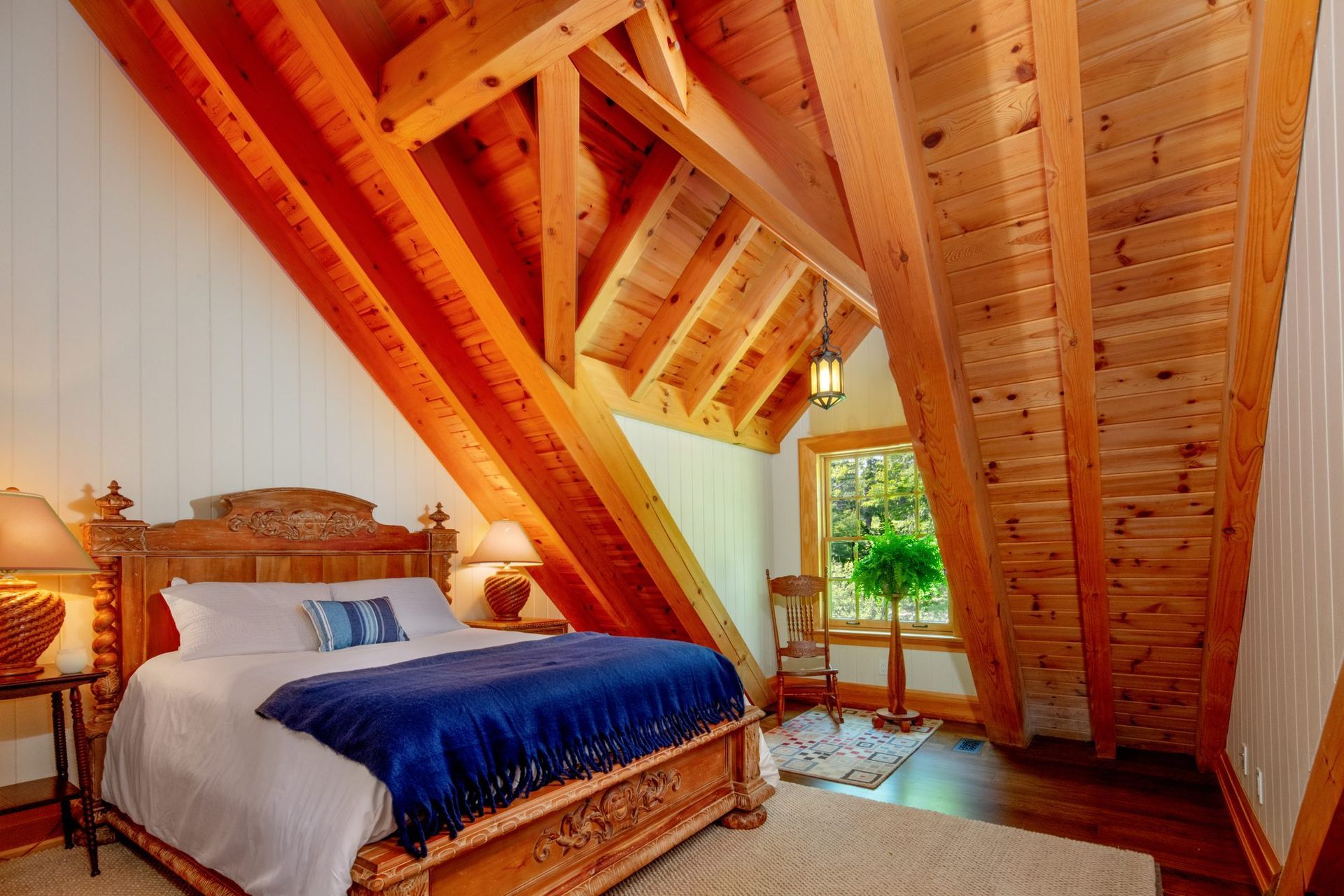 Cozy loft bedroom with slanted wooden ceiling, blue bedspread, and a window overlooking greenery