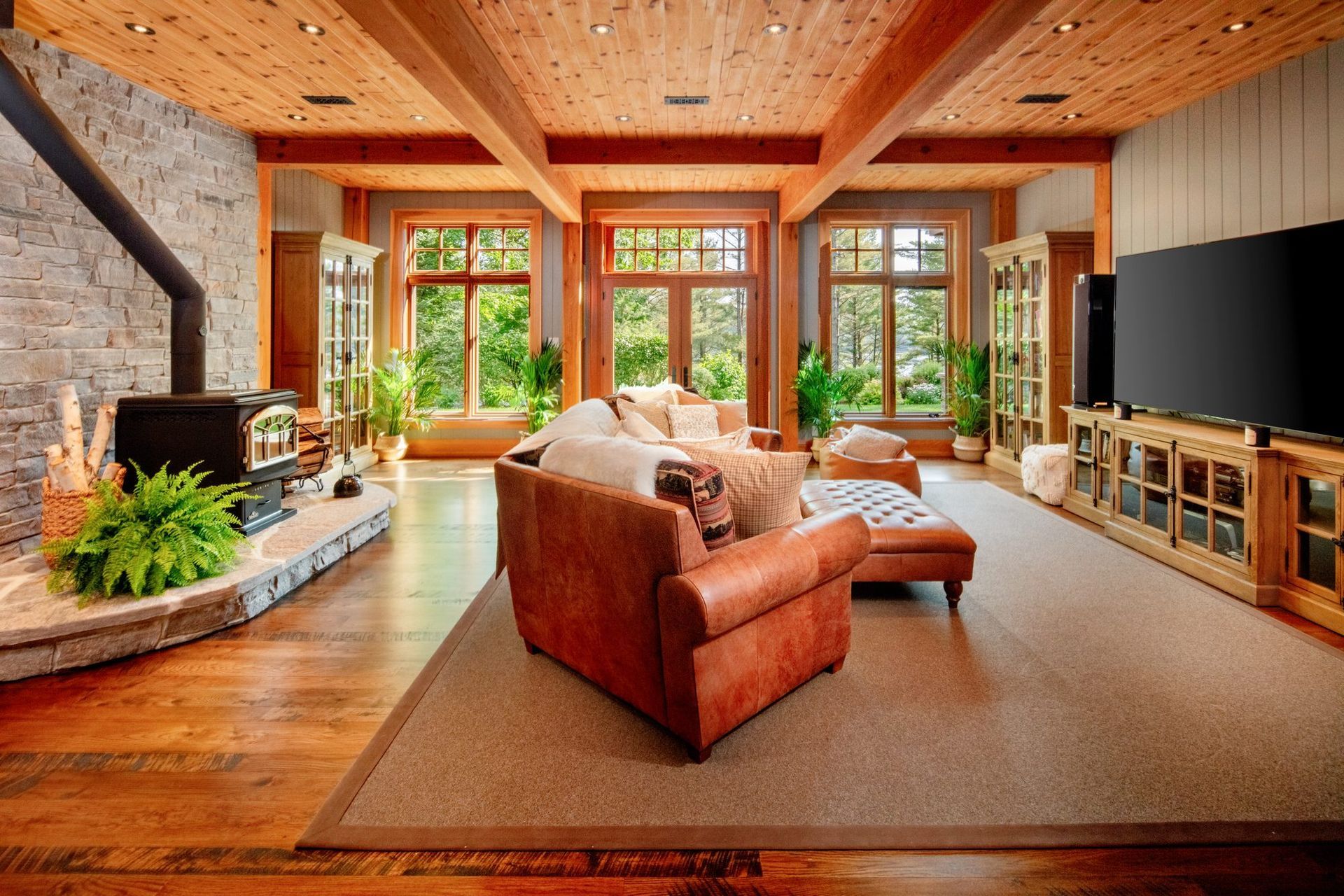 Modern living room with stone fireplace, brown sofa, large windows, wood ceiling, and TV console