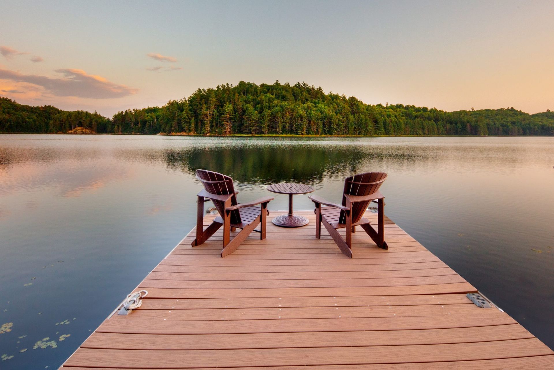 Wooden dock with two chairs facing a calm lake at sunset, with a forested island in the distance