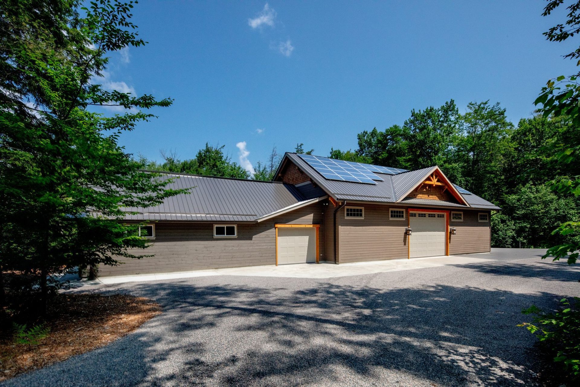 Modern house with gray roof and wooden garage doors, surrounded by trees and gravel driveway
