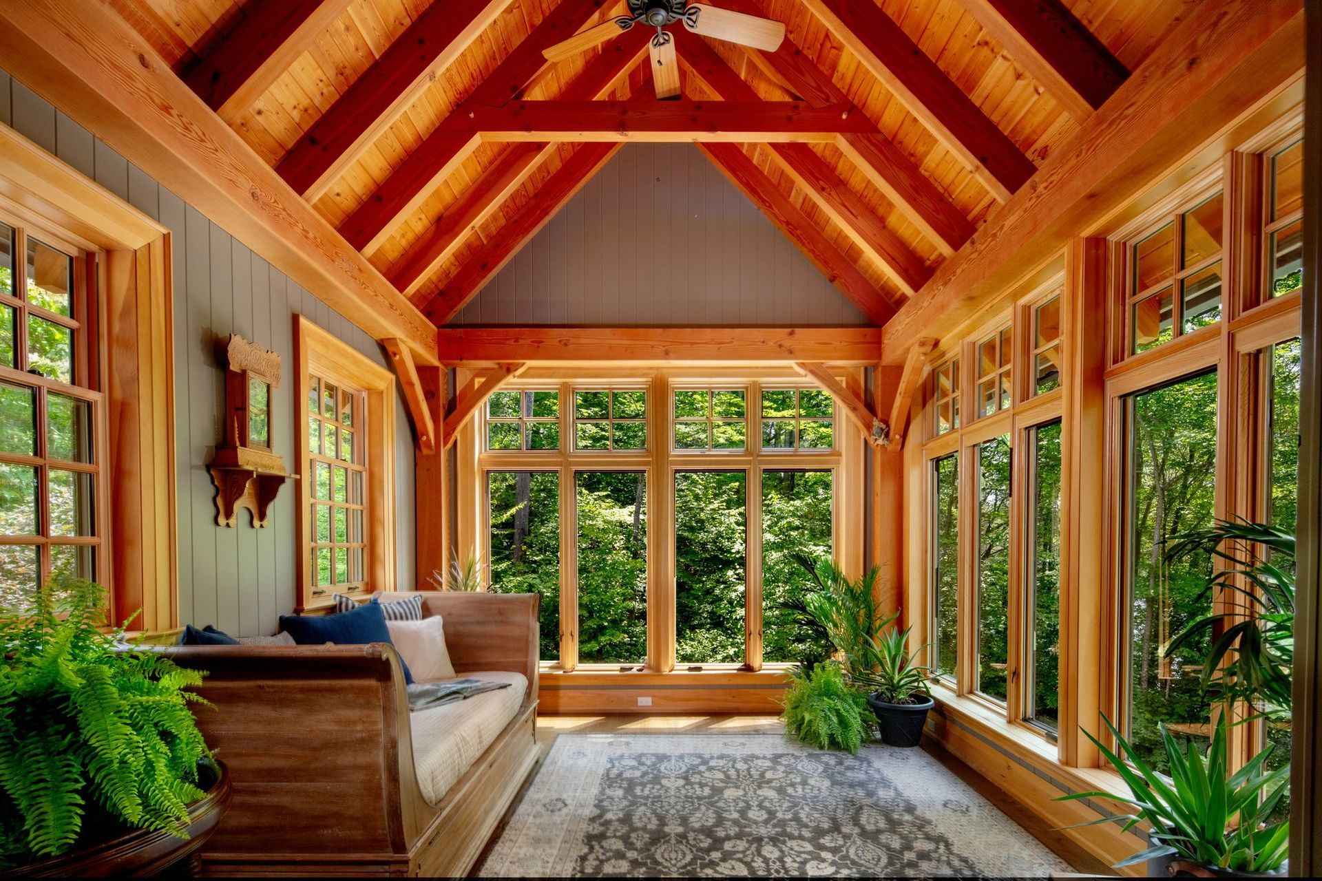 Sunlit wooden sunroom with vaulted beams, large windows, sofa, and lush indoor plants