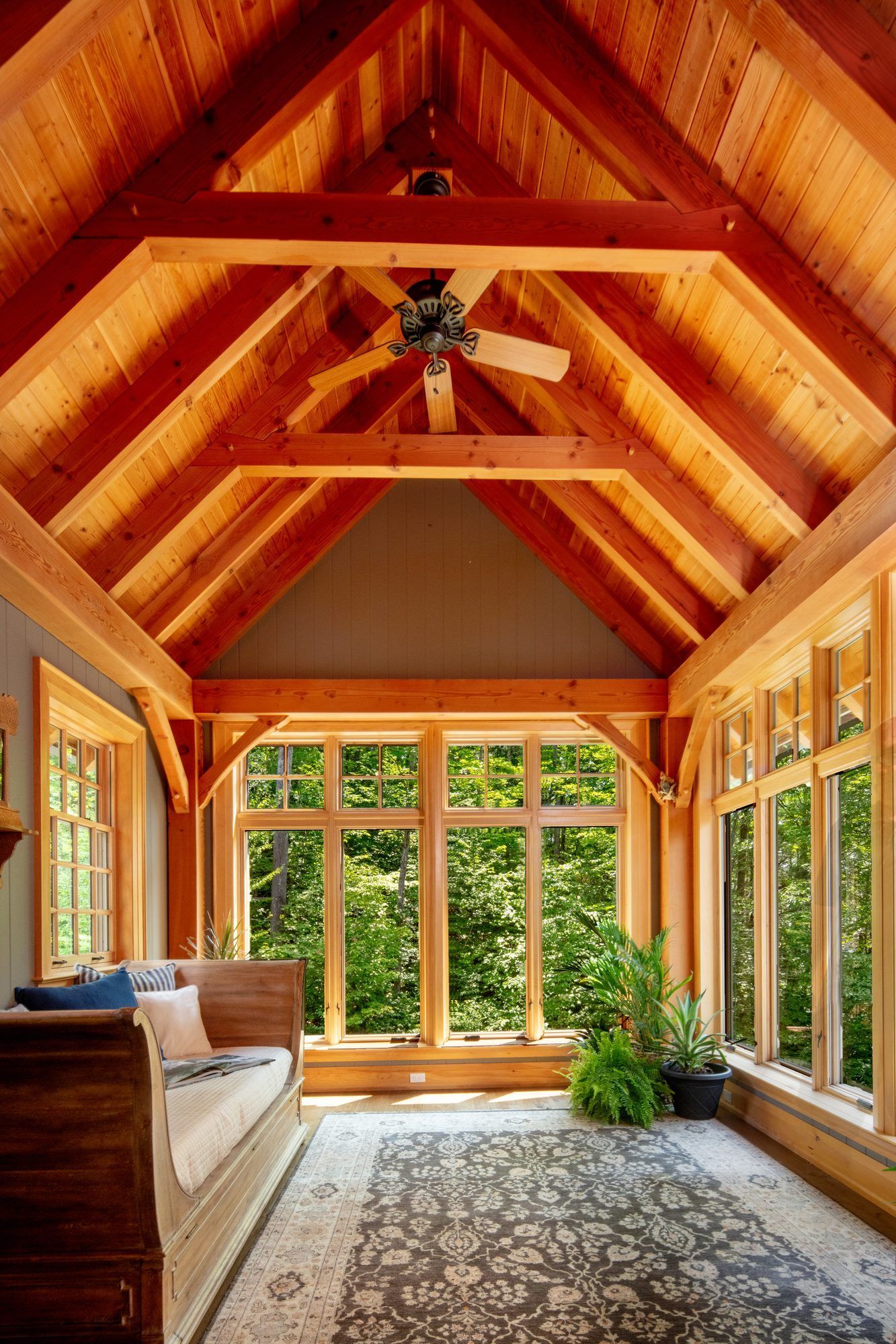 Wooden sunroom with vaulted ceiling, large windows, sofa, rug, and potted plant overlooking greenery