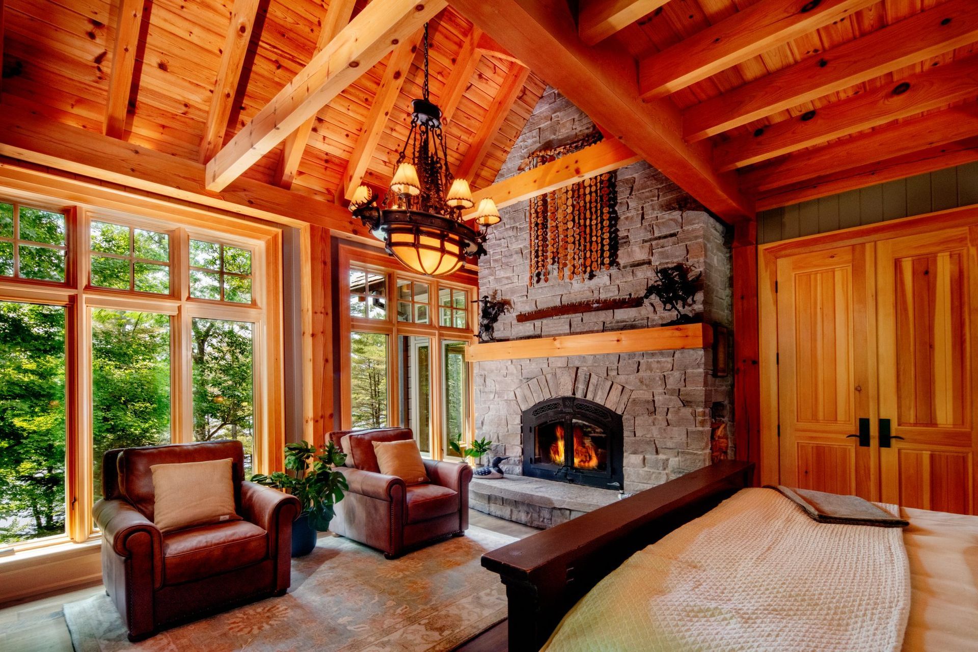Rustic cabin bedroom with stone fireplace, wood beams, large windows, and a bed by a wooden door