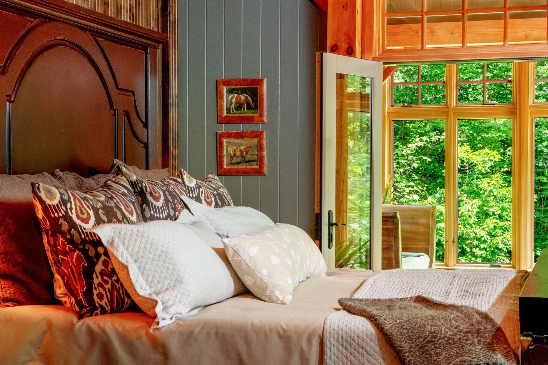 Bedroom with a wooden bed, patterned pillows, and large windows open to a green garden view.