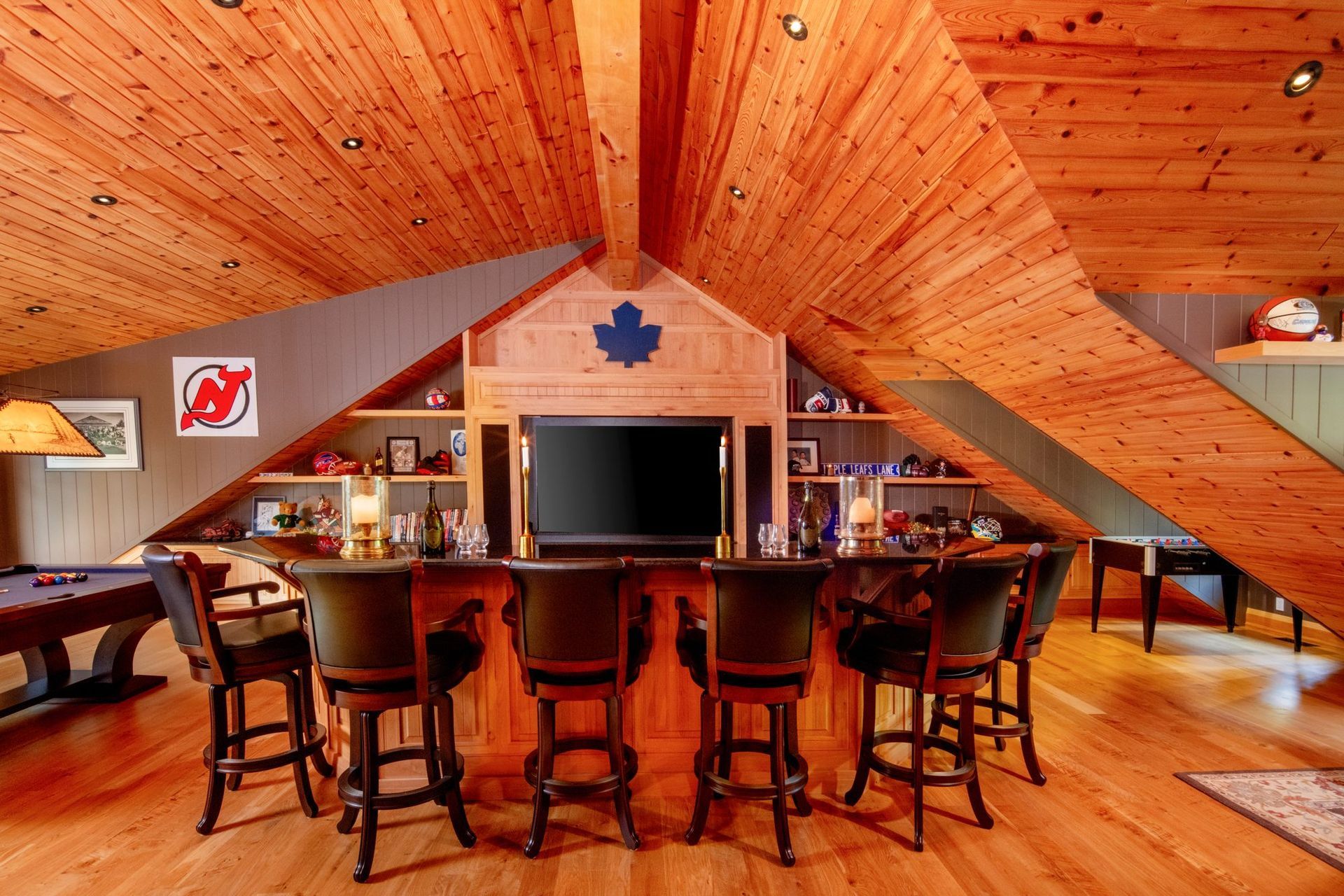 Rustic wood-paneled bar with stools, dining tables, and a large TV in a cozy attic-style room