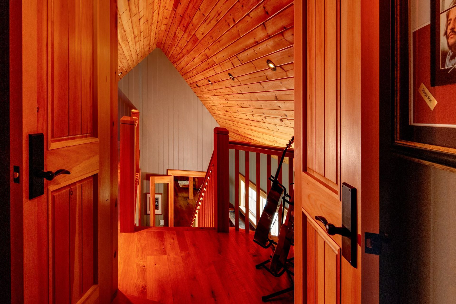 Warm-lit wooden hallway with open doors, a railing, and a sloped ceiling leading to stairs.