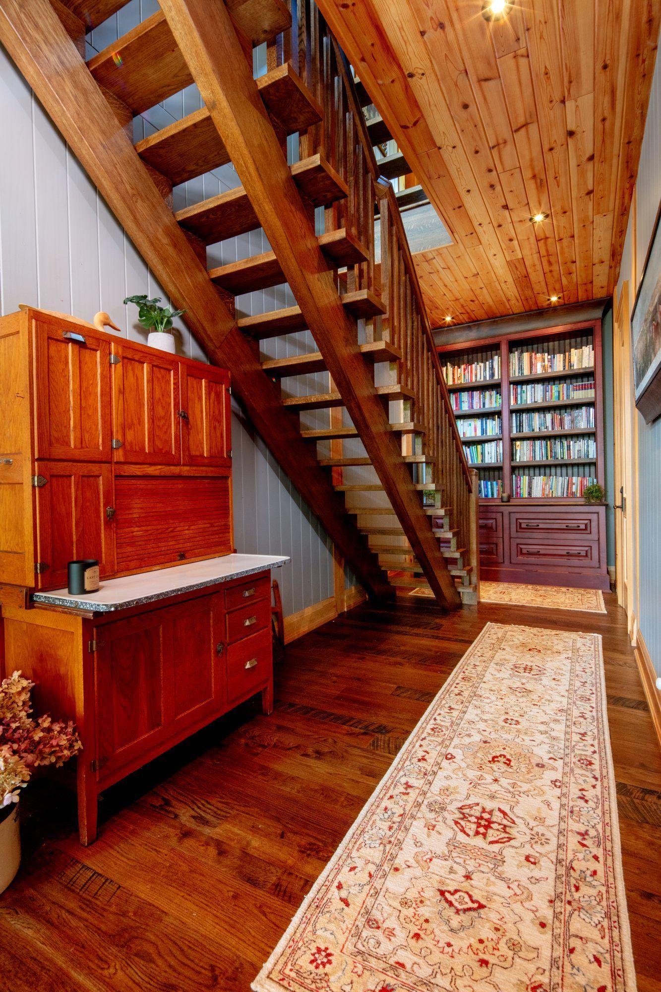 Narrow wooden hallway with a staircase, built-in bookshelves, cabinets, and a patterned runner rug.