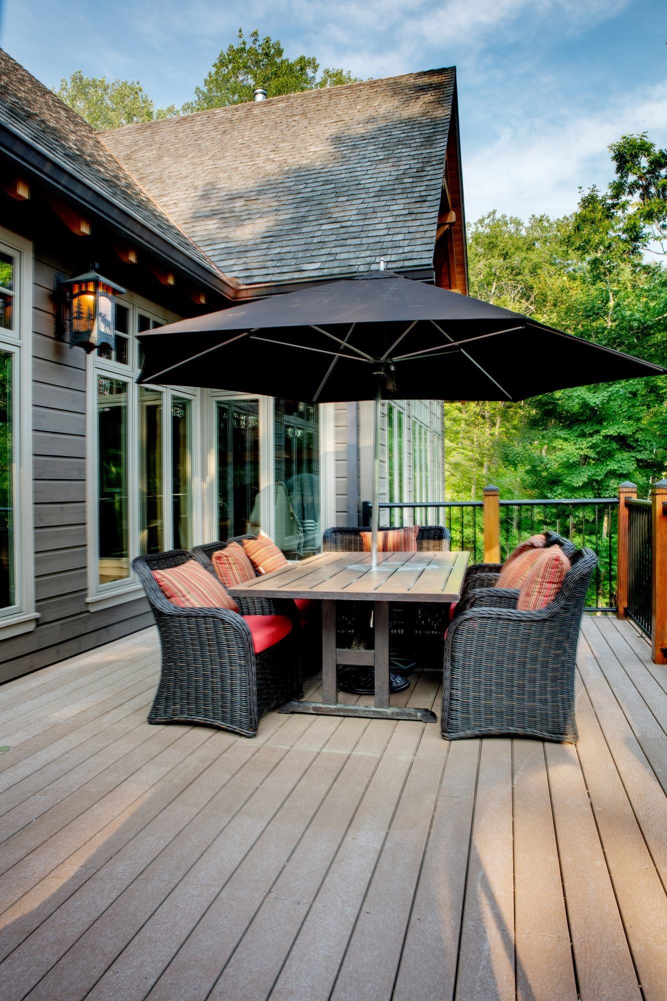 Outdoor deck with wicker chairs, dining table, and large black umbrella beside a house and trees.