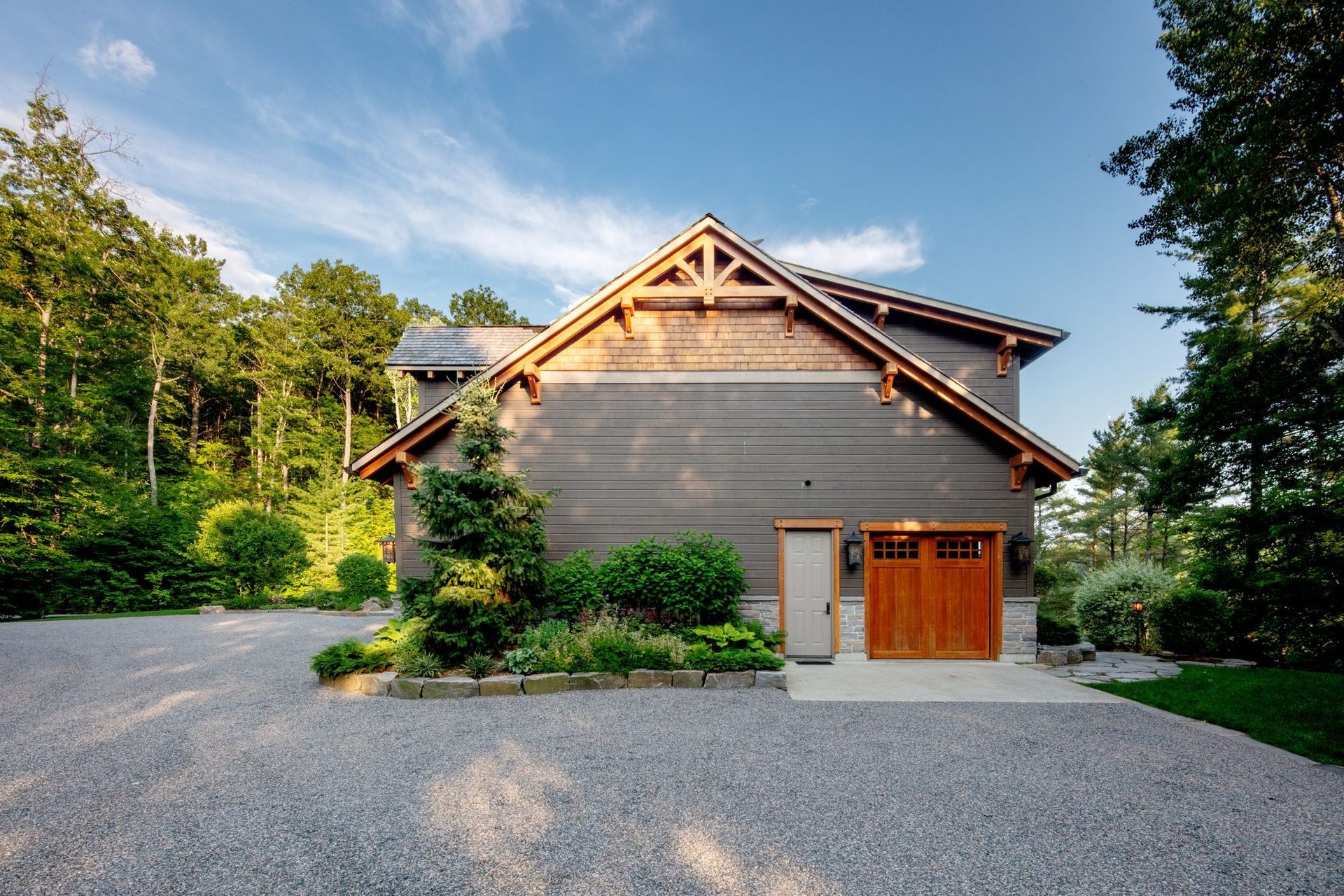 Wooden house with gravel driveway, surrounded by green trees under a clear blue sky