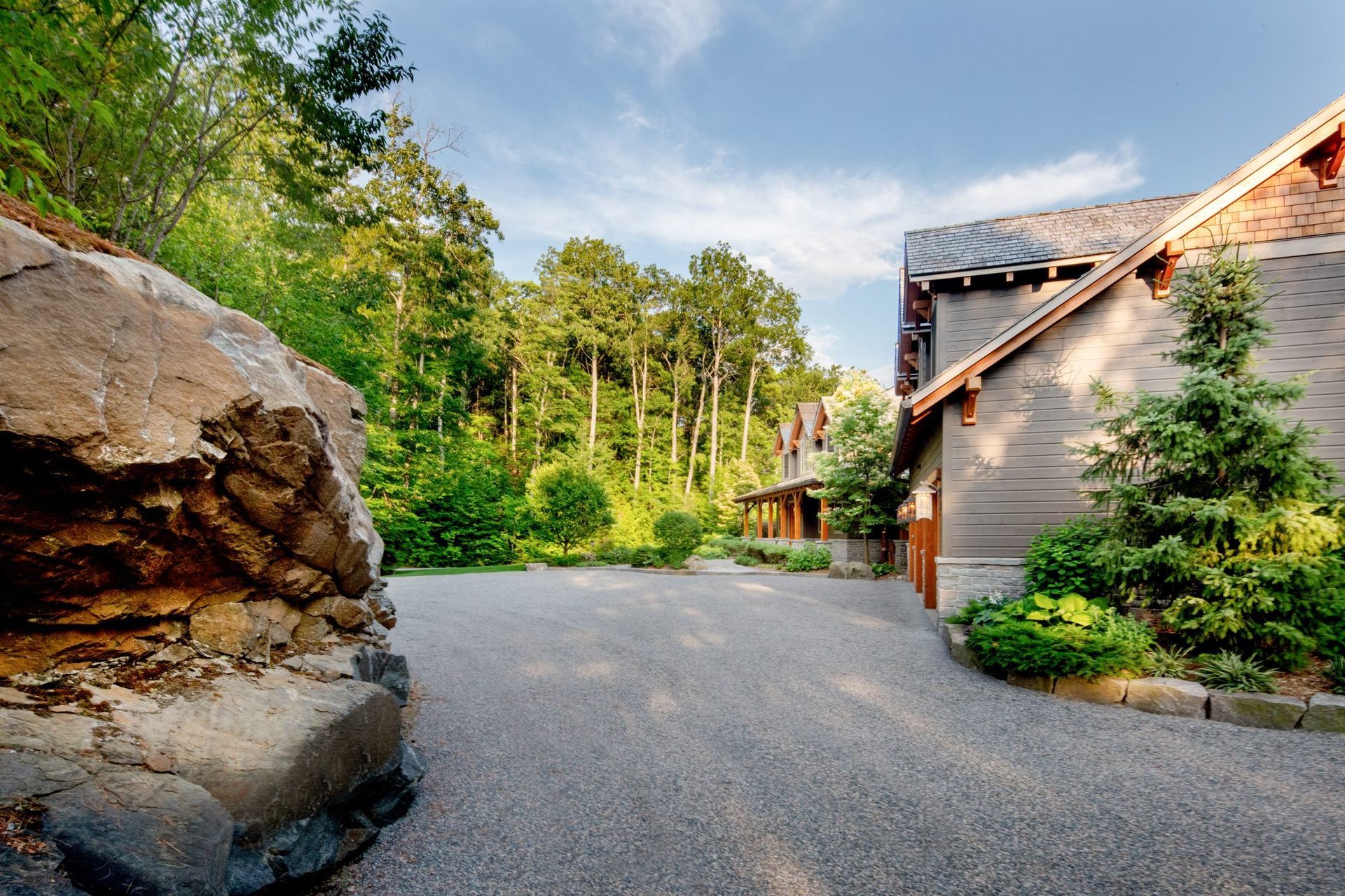 Gravel driveway beside a stone house and large rock, surrounded by trees under a blue sky.
