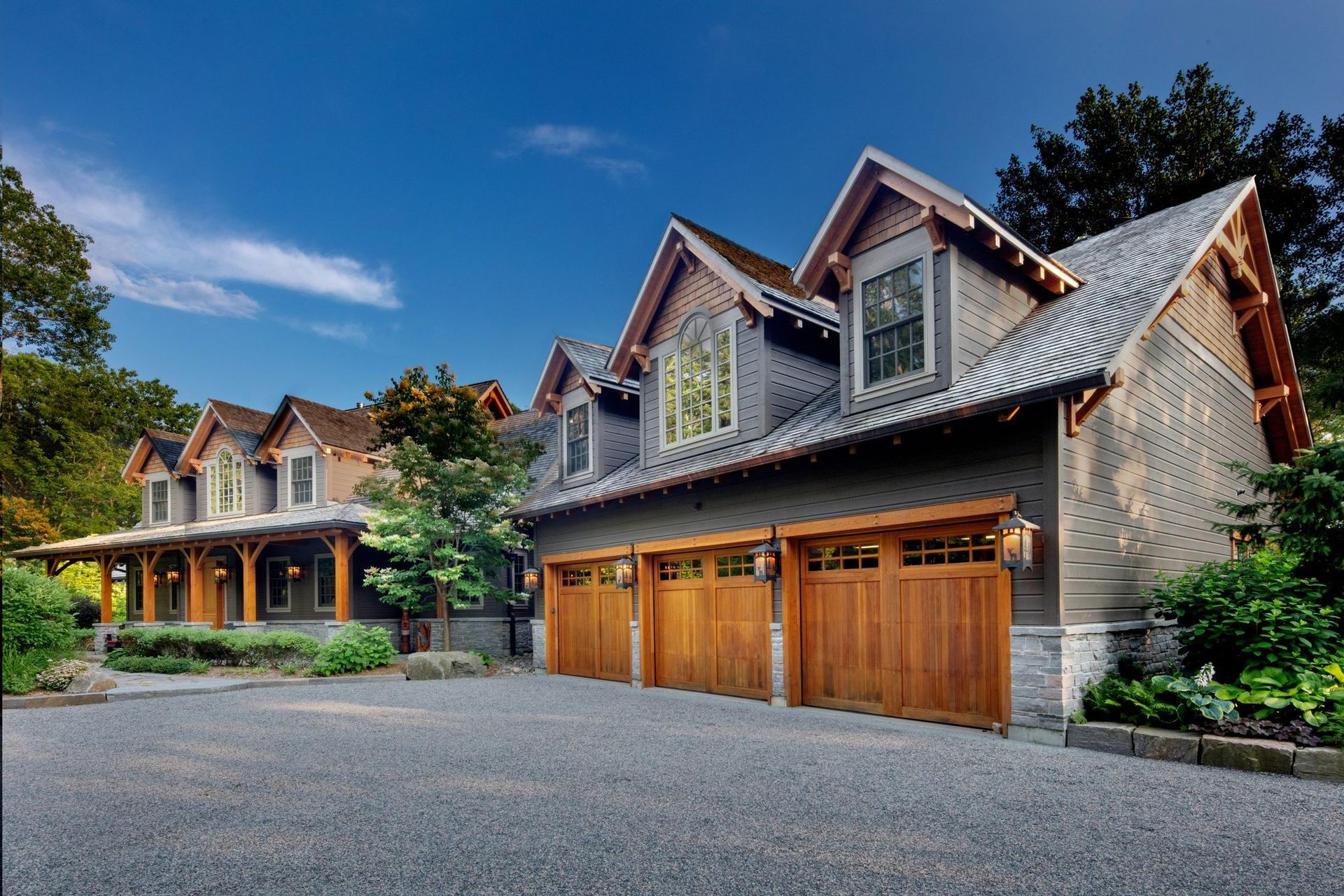 Stone house with wooden garage doors and dormer windows in a landscaped driveway under a blue sky