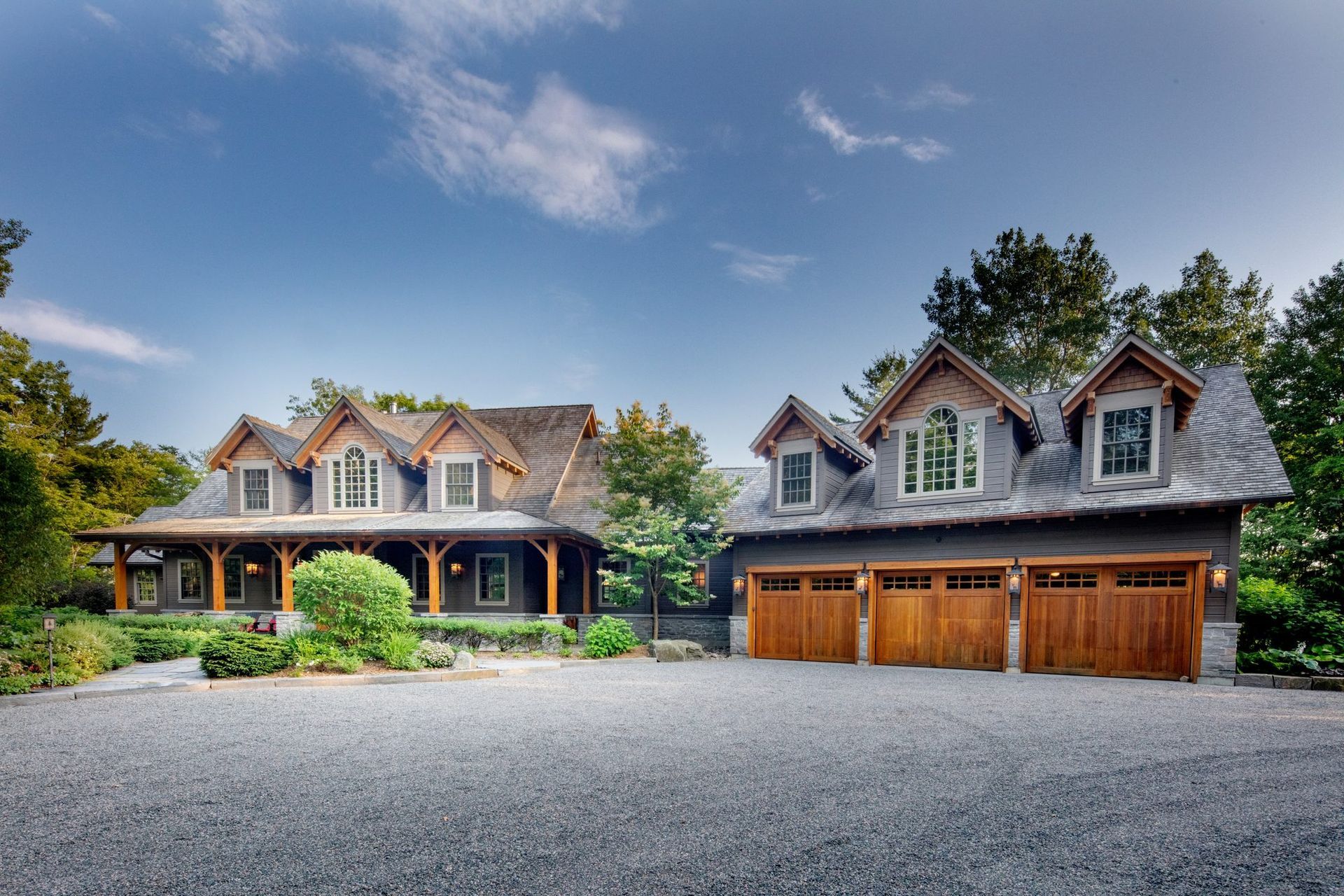 Large rustic house with a covered porch, stone garage, and gravel driveway under a blue sky