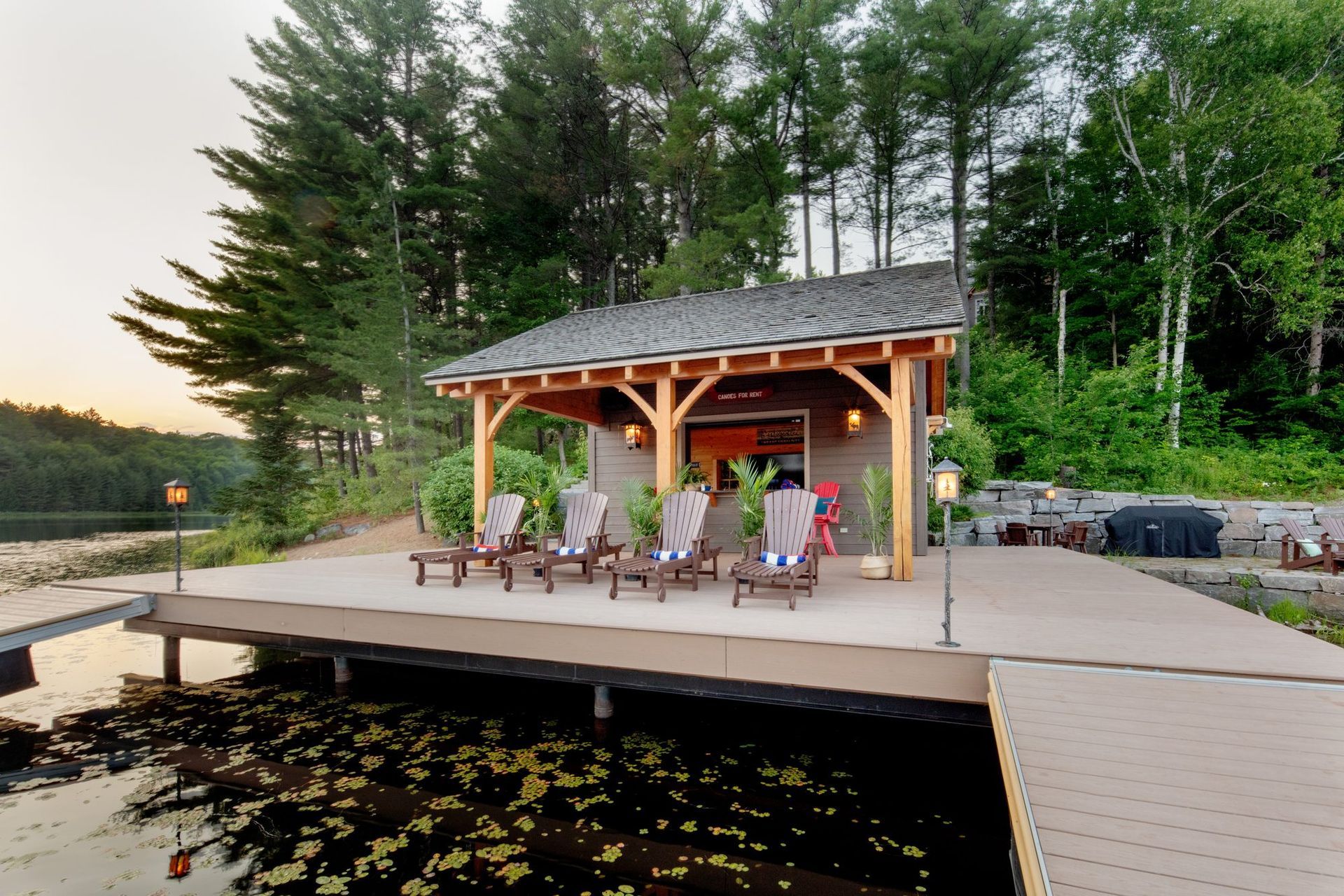 Lakeside pavilion with people seated on a wooden deck at sunset, surrounded by trees.