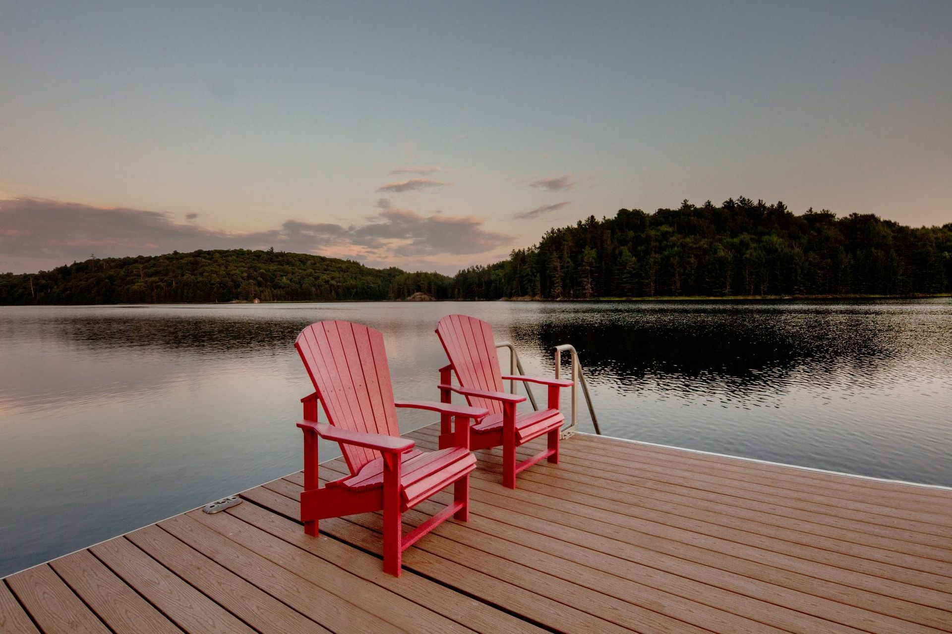 Two red Adirondack chairs on a wooden dock beside a calm lake at sunset