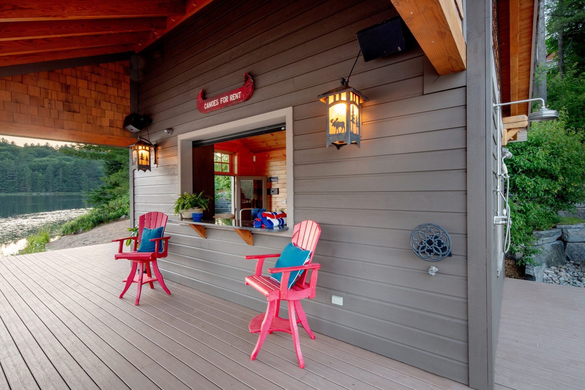 Covered porch with pink chairs and a lantern beside a rustic gray cabin window.