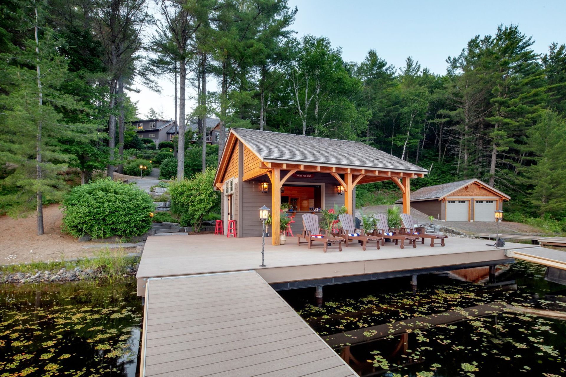 Lakeside cabin with covered deck and chairs at the end of a wooden dock, surrounded by trees.
