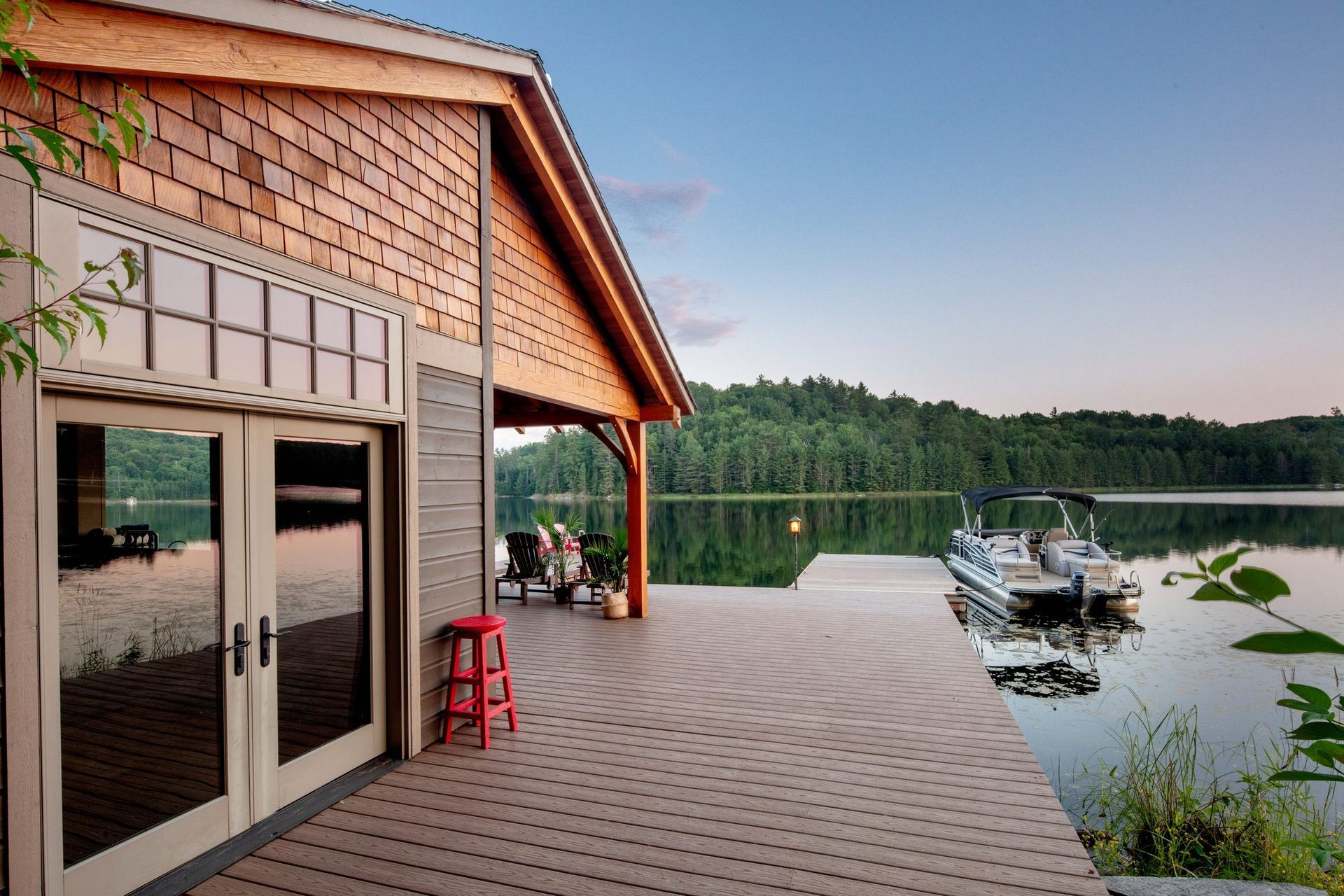 Lakeside cabin porch with docked boat, red stools, and forested shoreline at sunset