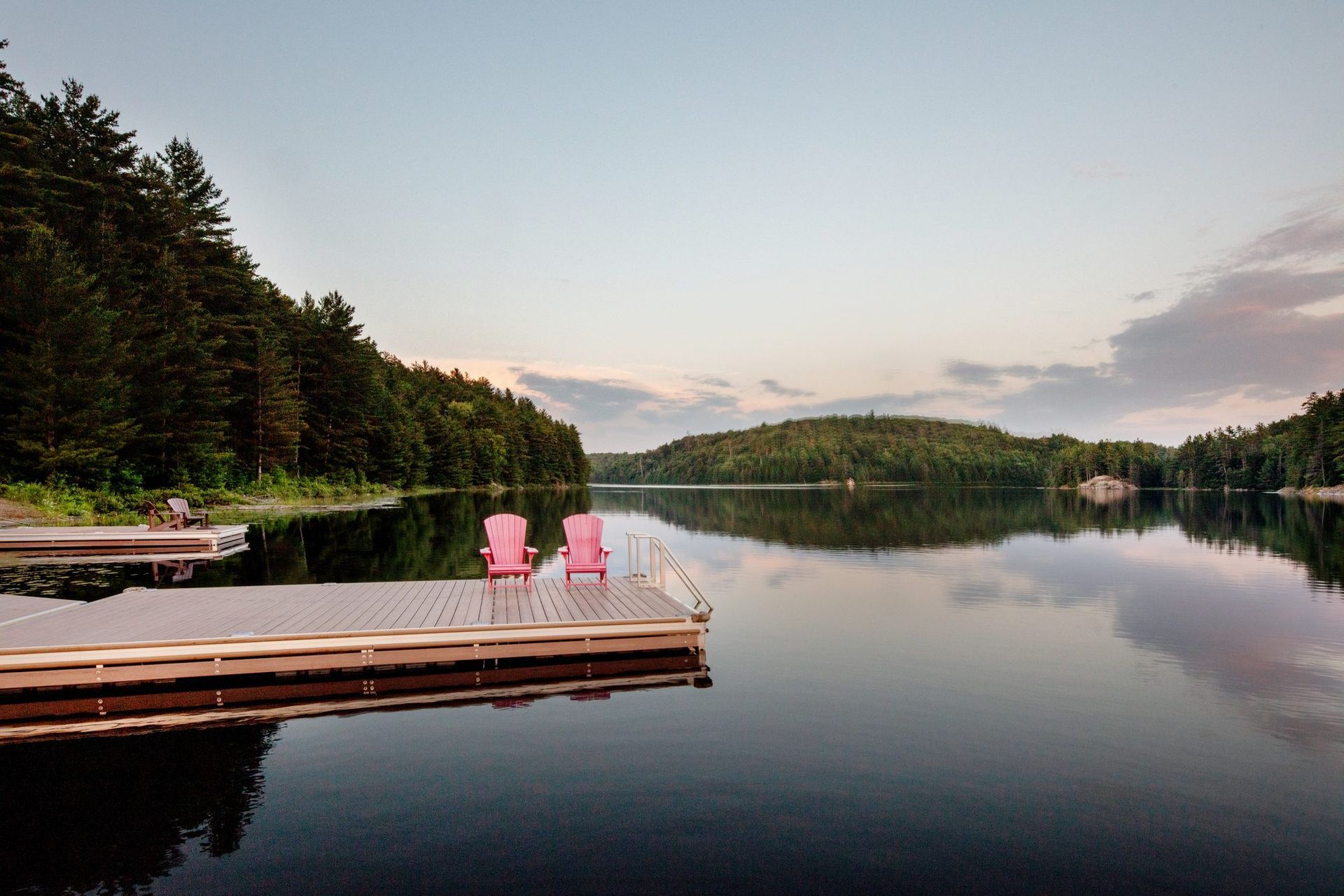 Wooden dock with pink chairs on a calm lake at dusk, surrounded by forest and reflections