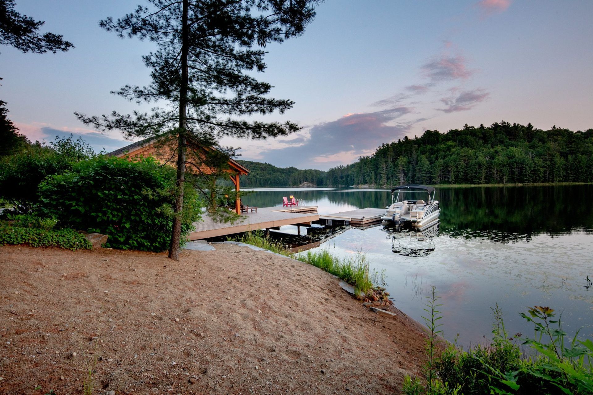 Quiet lakeside dock at dusk with boats, forested shoreline, and a sandy path in the foreground