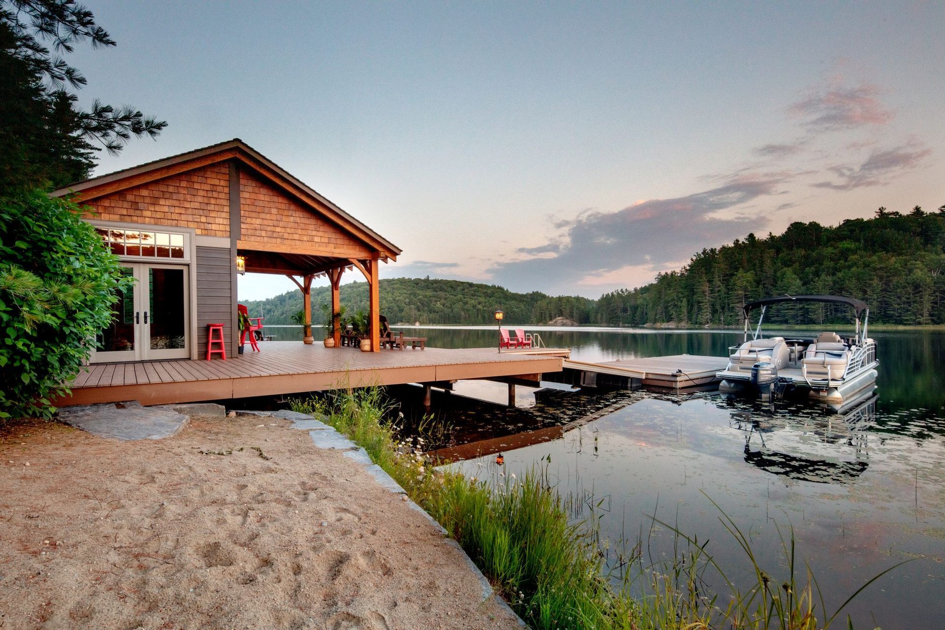 Lakeside cabin with dock at sunset, boats moored on calm water, and a sandy shore in the foreground
