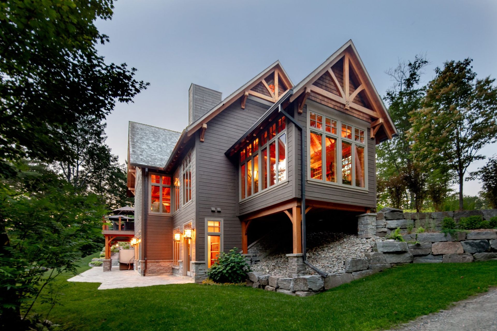 Stone-and-wood house with warm lights, large windows, and a sloped roof surrounded by trees and grass