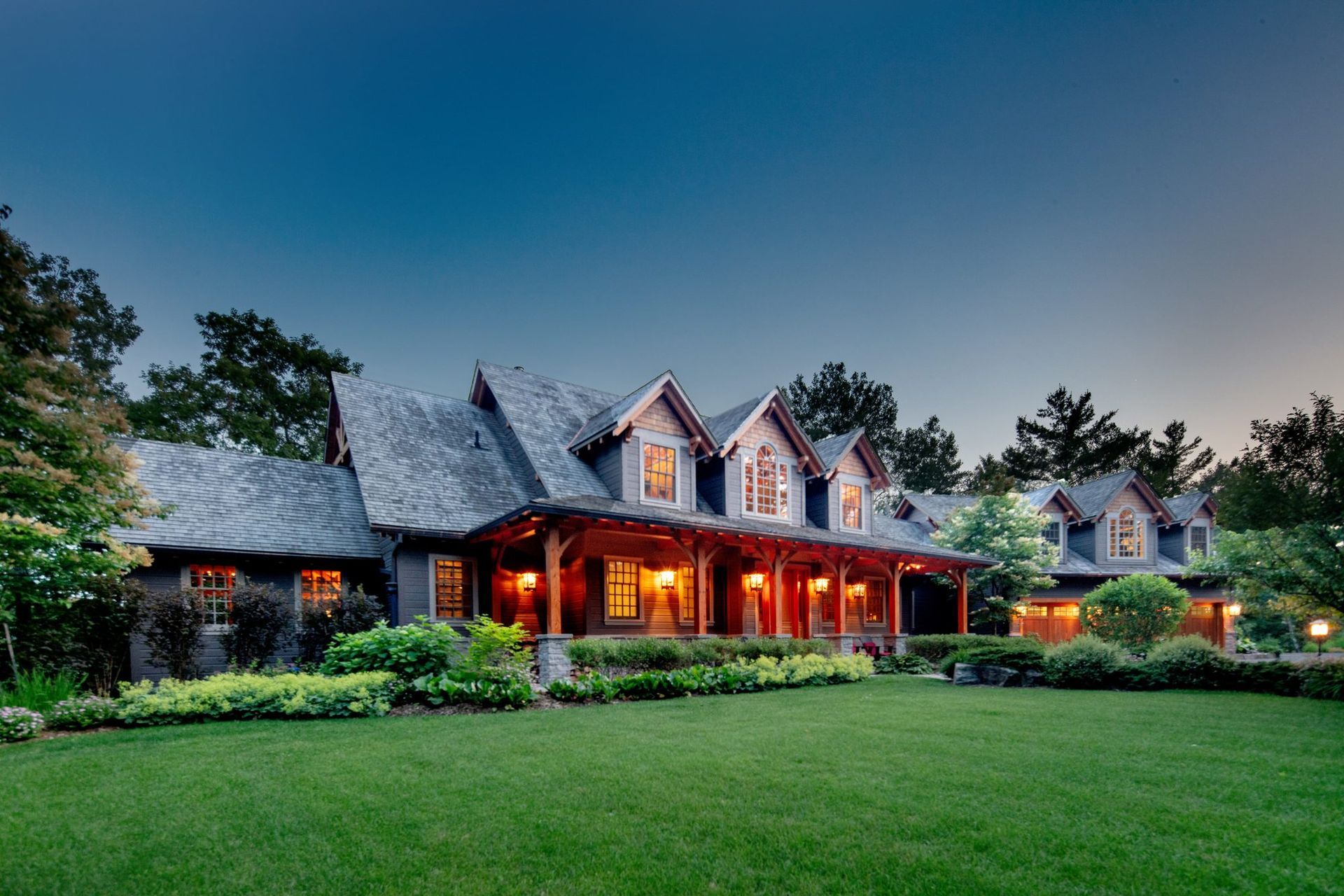Large stone house with warm porch lights at dusk, surrounded by a manicured lawn and trees.