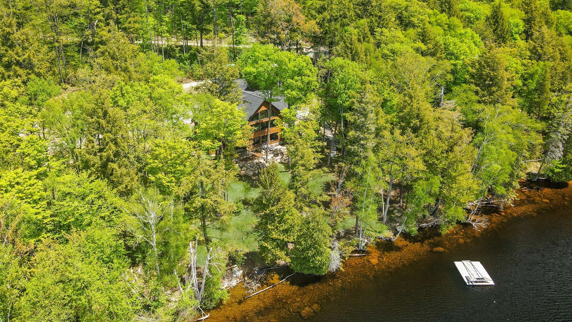 Aerial view of a forested lakeshore with a small cabin and a dock on dark water