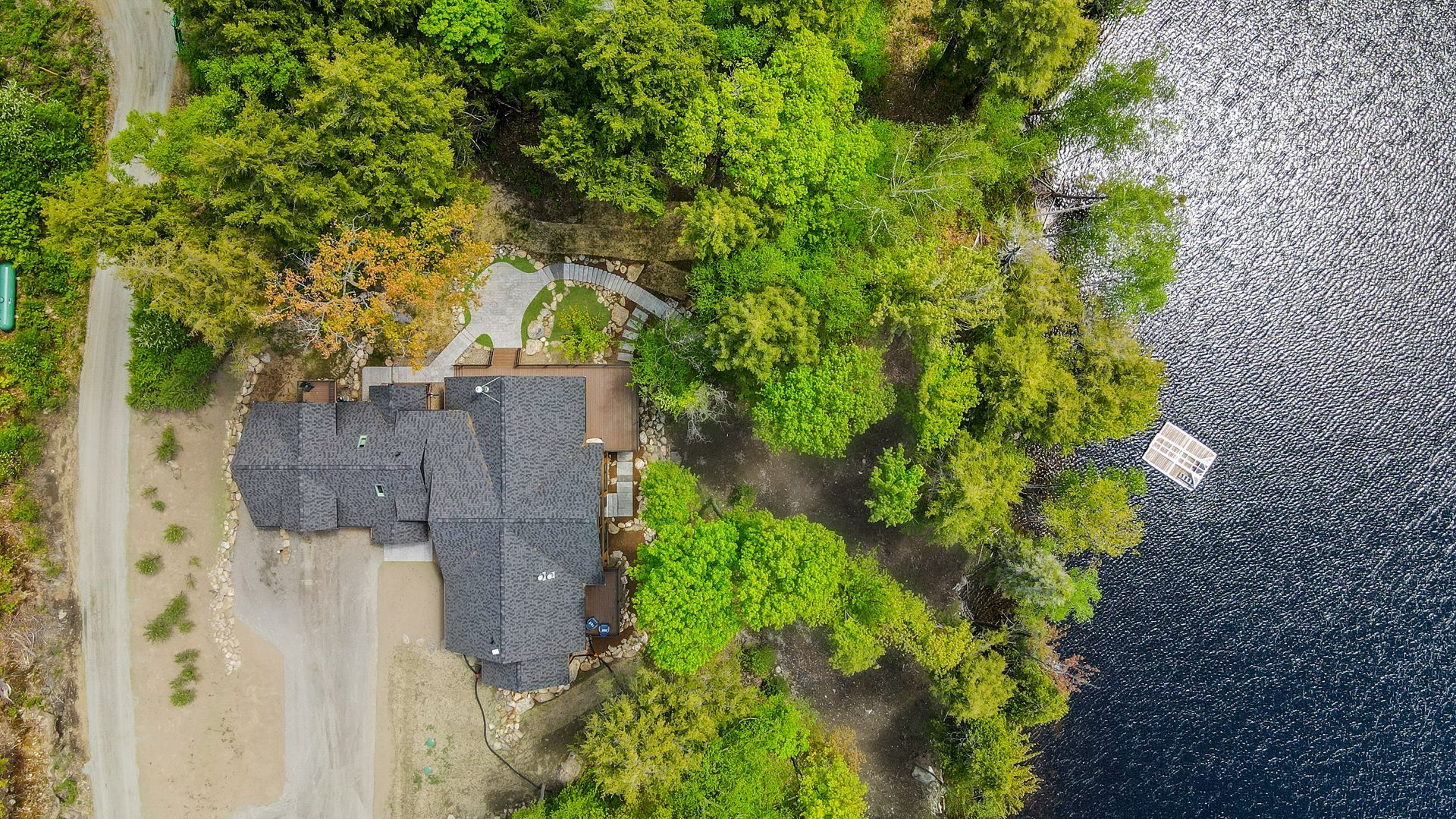 Aerial view of a lakeside house surrounded by trees beside a gravel shore and dark water.