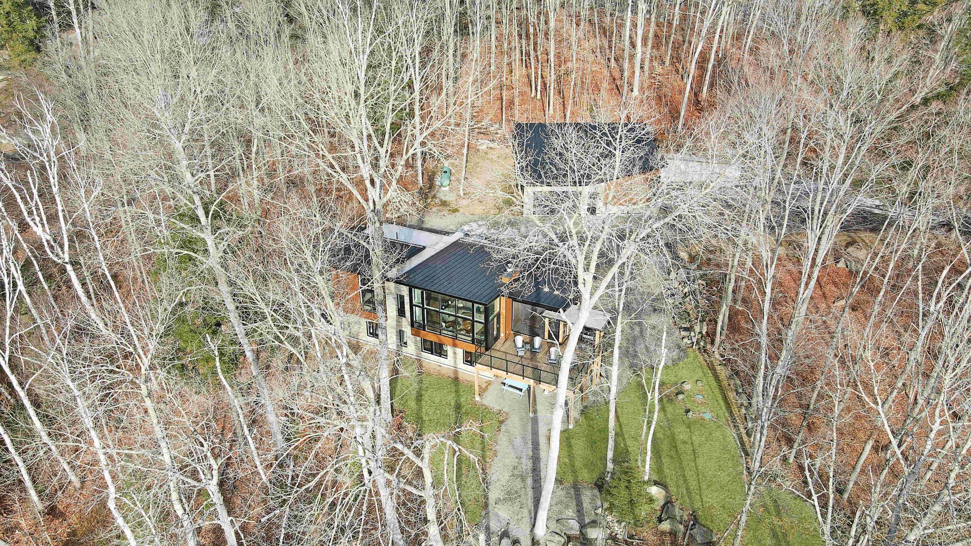 Aerial view of a dark-roofed cabin surrounded by leafless trees and green grass.
