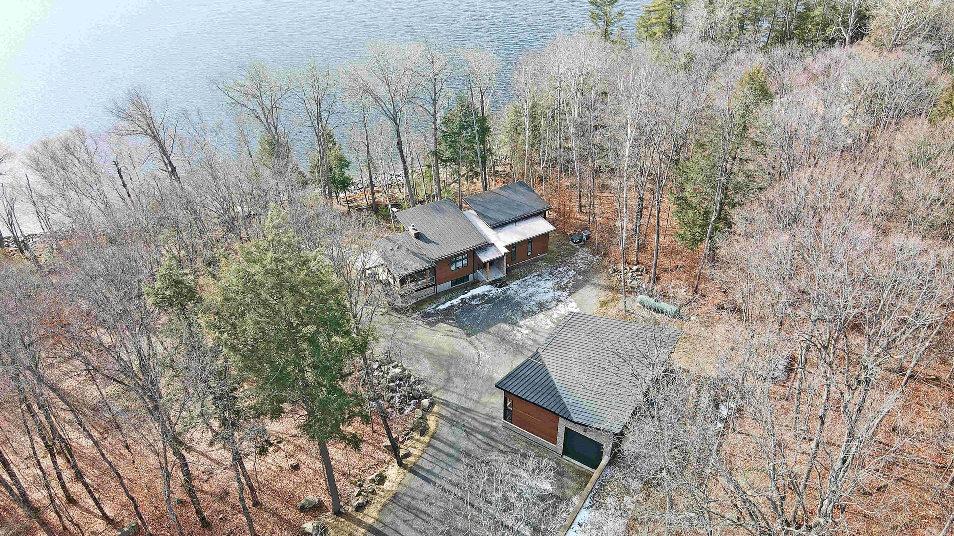 Aerial view of two cabins in a snowy wooded area with bare trees and a misty lake in the background