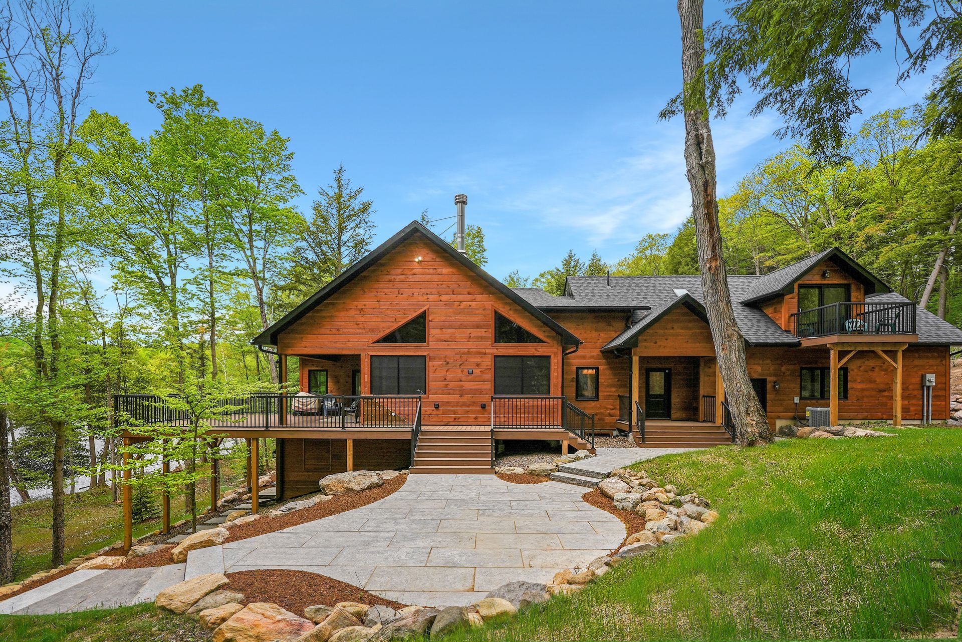 Rustic log cabin with stone chimney and large windows, surrounded by trees and a stone driveway