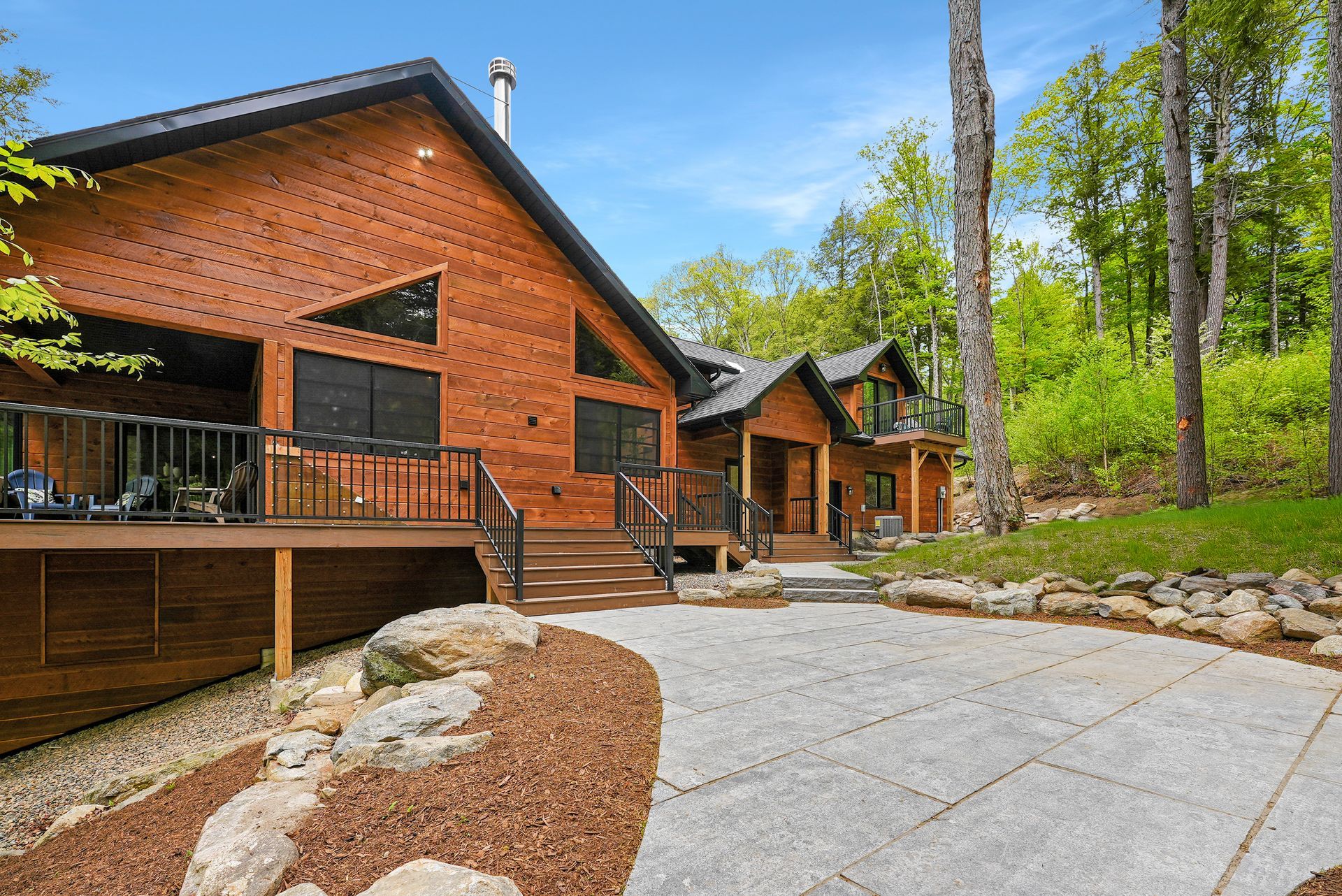 Modern red brick cabin with wooden deck beside a paved driveway in a wooded setting