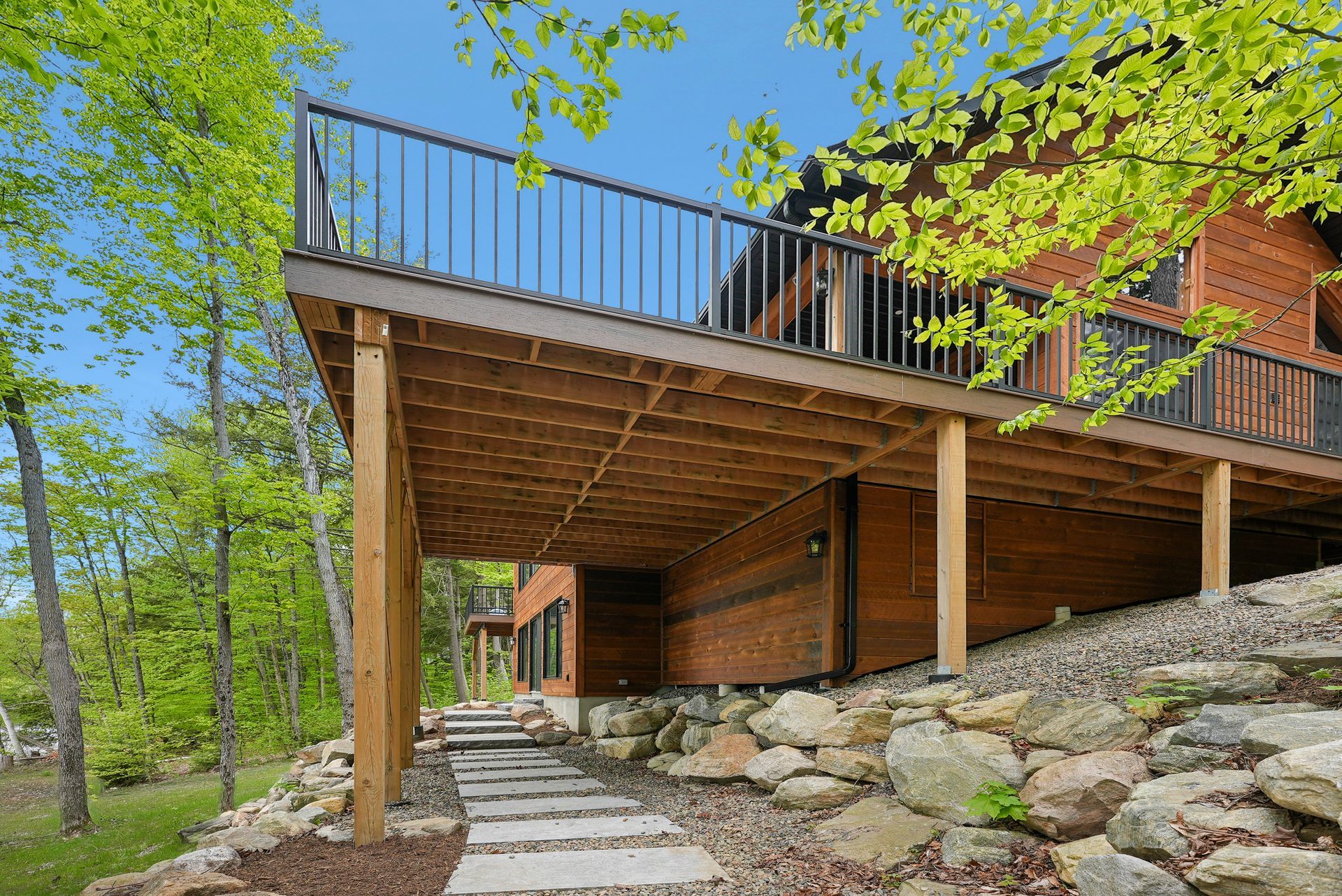 Modern wooden deck on stilts with stone landscaping and a paved path in a wooded area