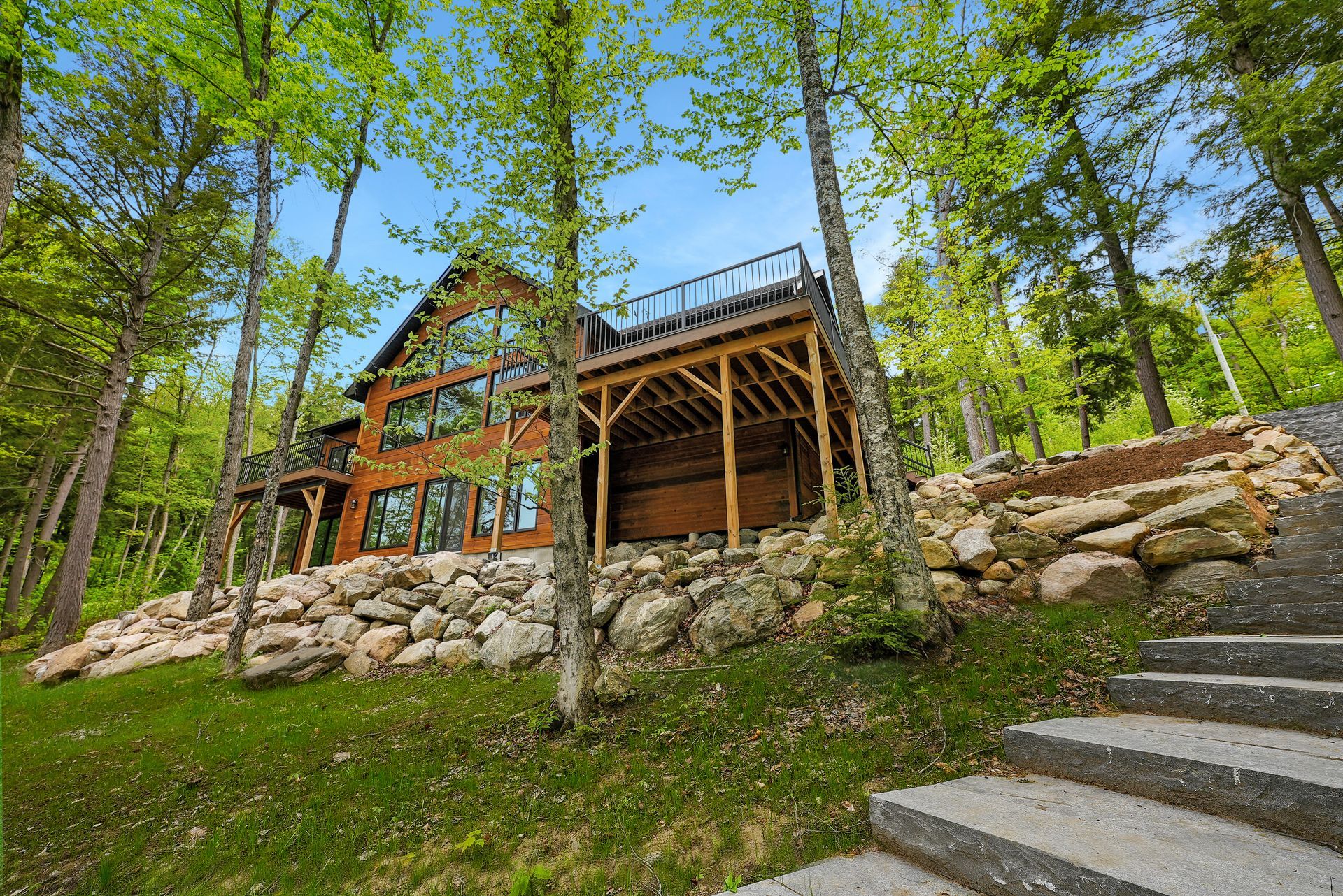 Cabin in the woods with stone steps, wooden deck, and tall trees on a sunny hillside