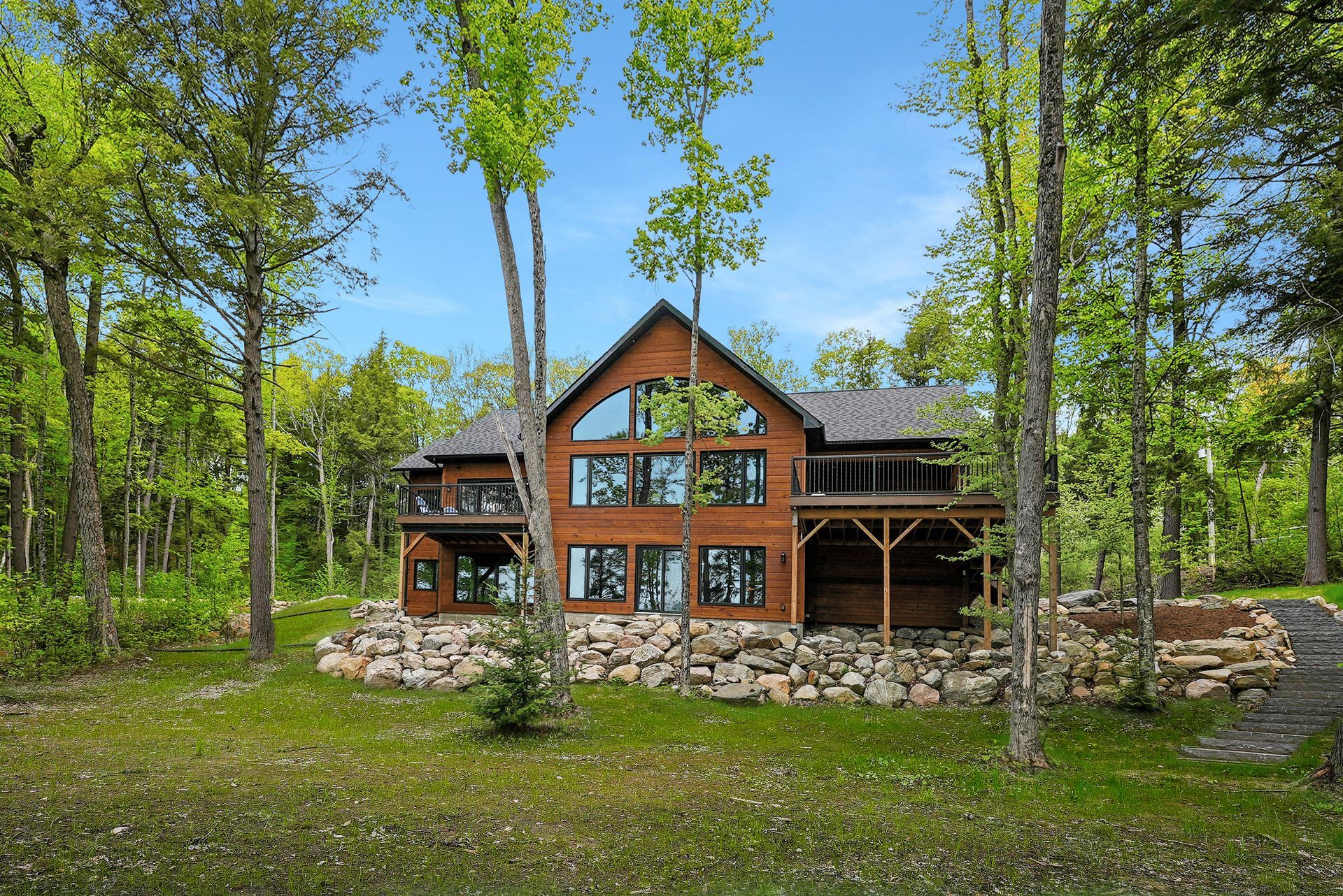 Rustic log cabin with stone foundation and large windows, surrounded by trees and greenery under a blue sky