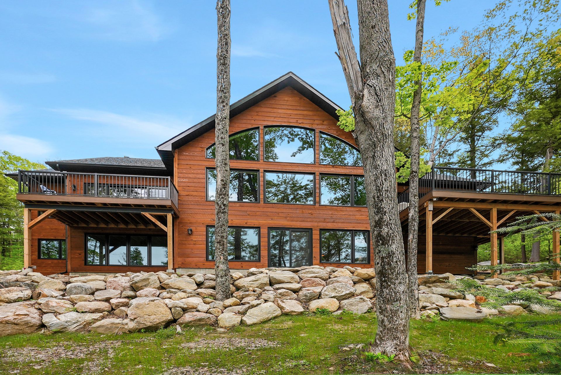 Rustic log cabin with large windows and stone retaining wall amid trees and greenery