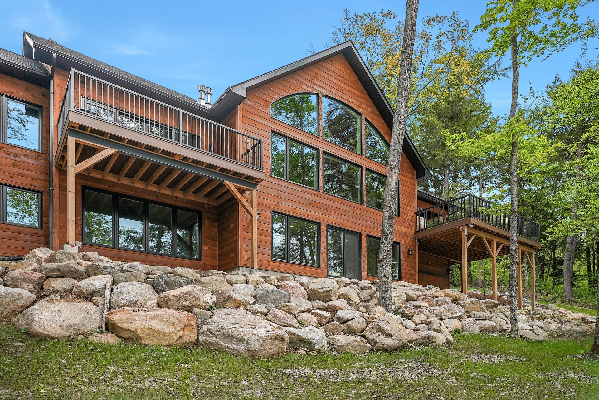 Rustic red-brick cabin with large windows, wooden deck, and stone retaining wall in a wooded setting