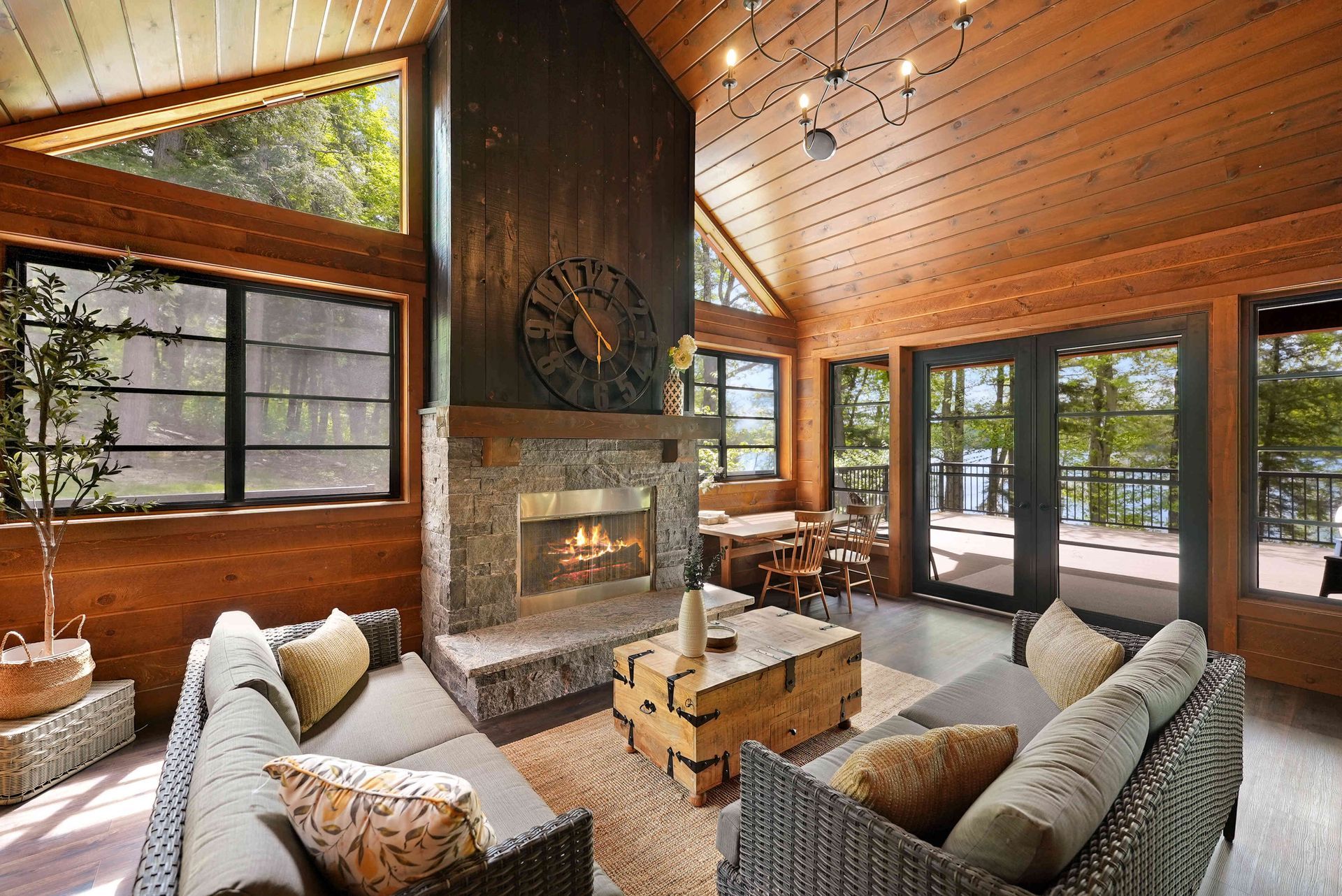 Cozy living room with a stone fireplace, vaulted wood ceiling, and large windows overlooking trees