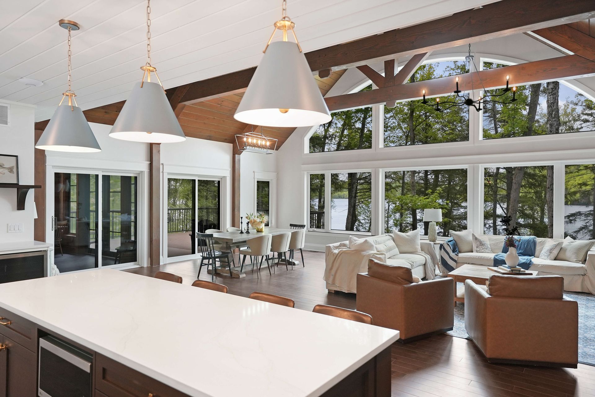 Bright open-concept living room with white vaulted ceilings, large windows, and tan seating near a kitchen island.