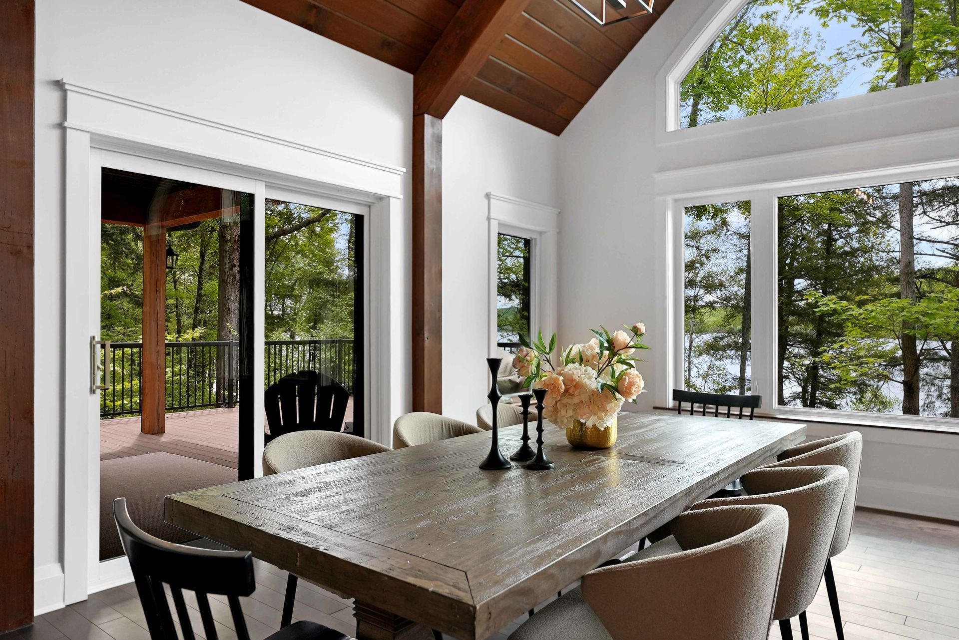 Bright dining room with a long table, beige chairs, and large windows overlooking trees and a deck.