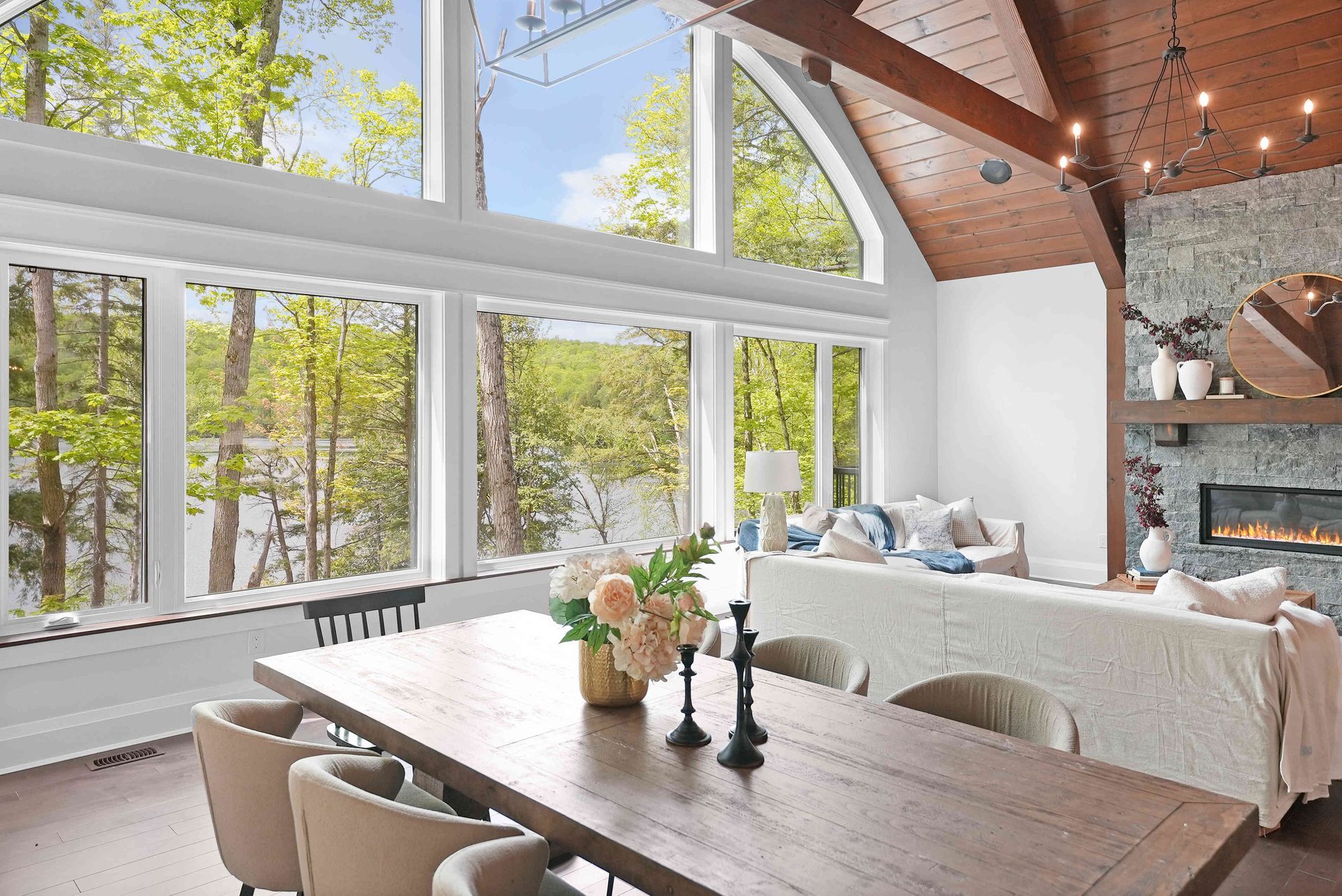 Bright open dining and living room with large windows, wood beams, white decor, and a lake view