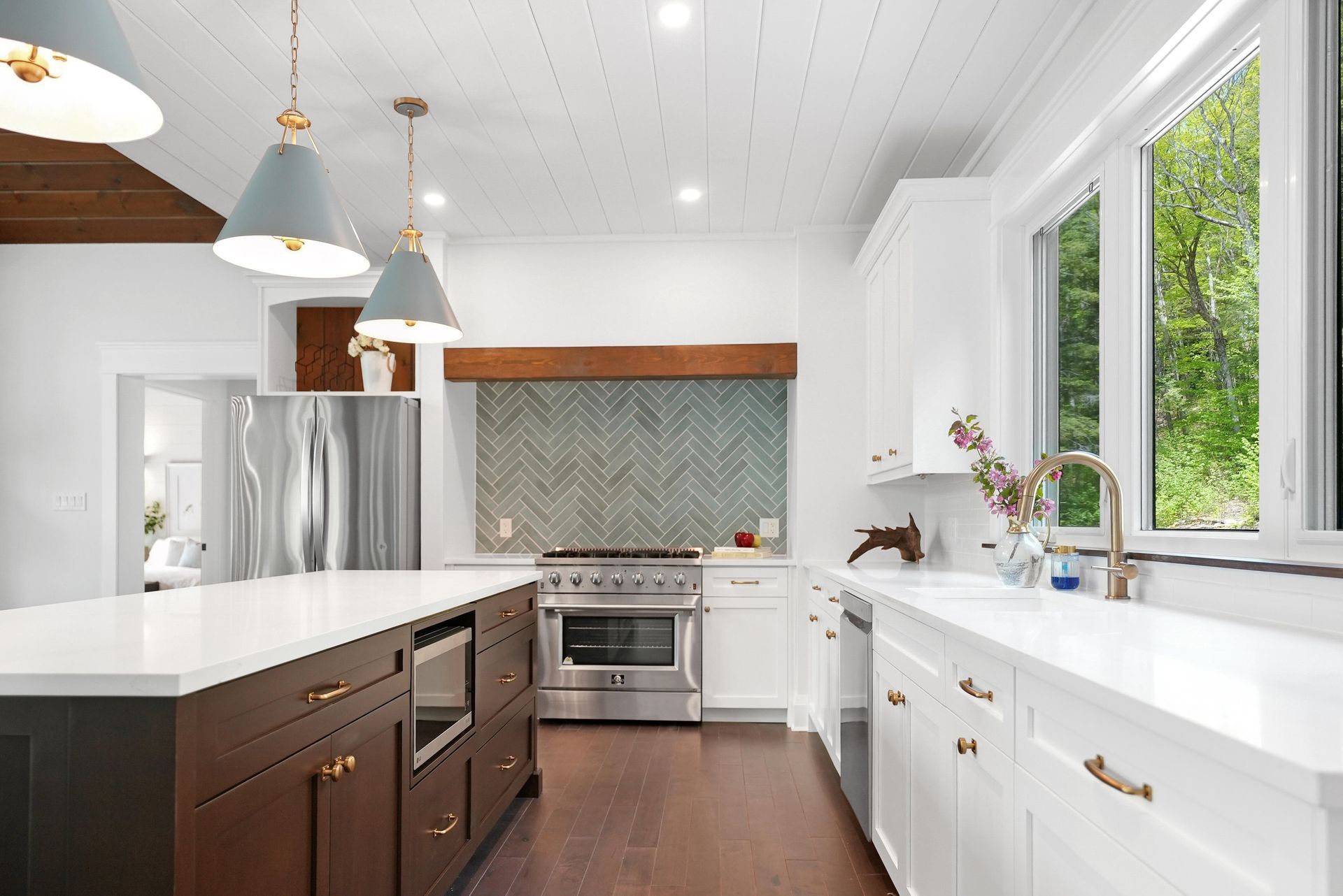 Modern white kitchen with dark island, stainless steel appliances, and large windows overlooking greenery