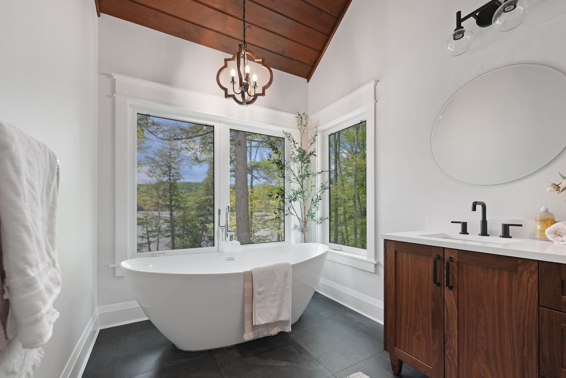 Bright bathroom with a freestanding tub, wood vanity, round mirror, and large windows overlooking trees.