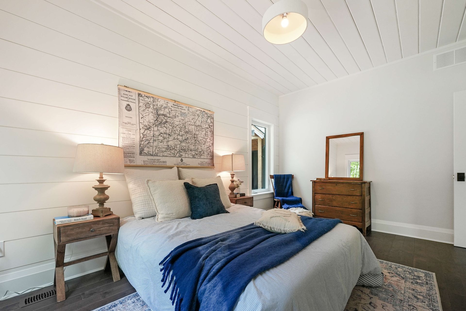 Bright bedroom with a blue bedspread, twin nightstands, dresser, mirror, and white vaulted ceiling.